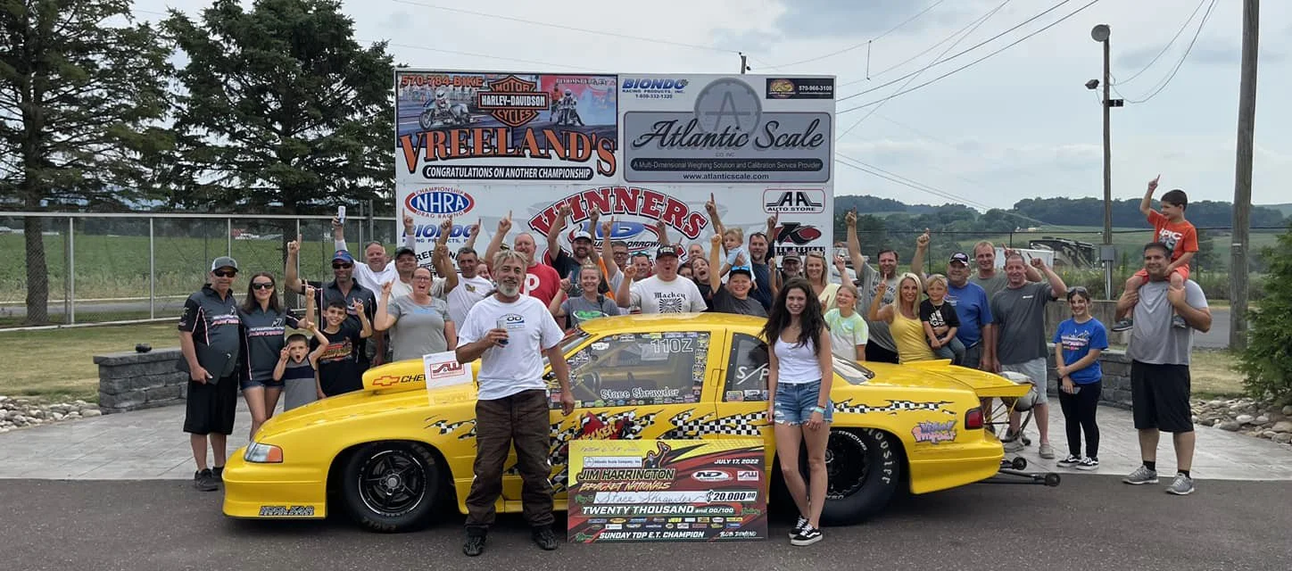 Group of people celebrating around a yellow race car with a large check and trophy in front, at a race track or event.
