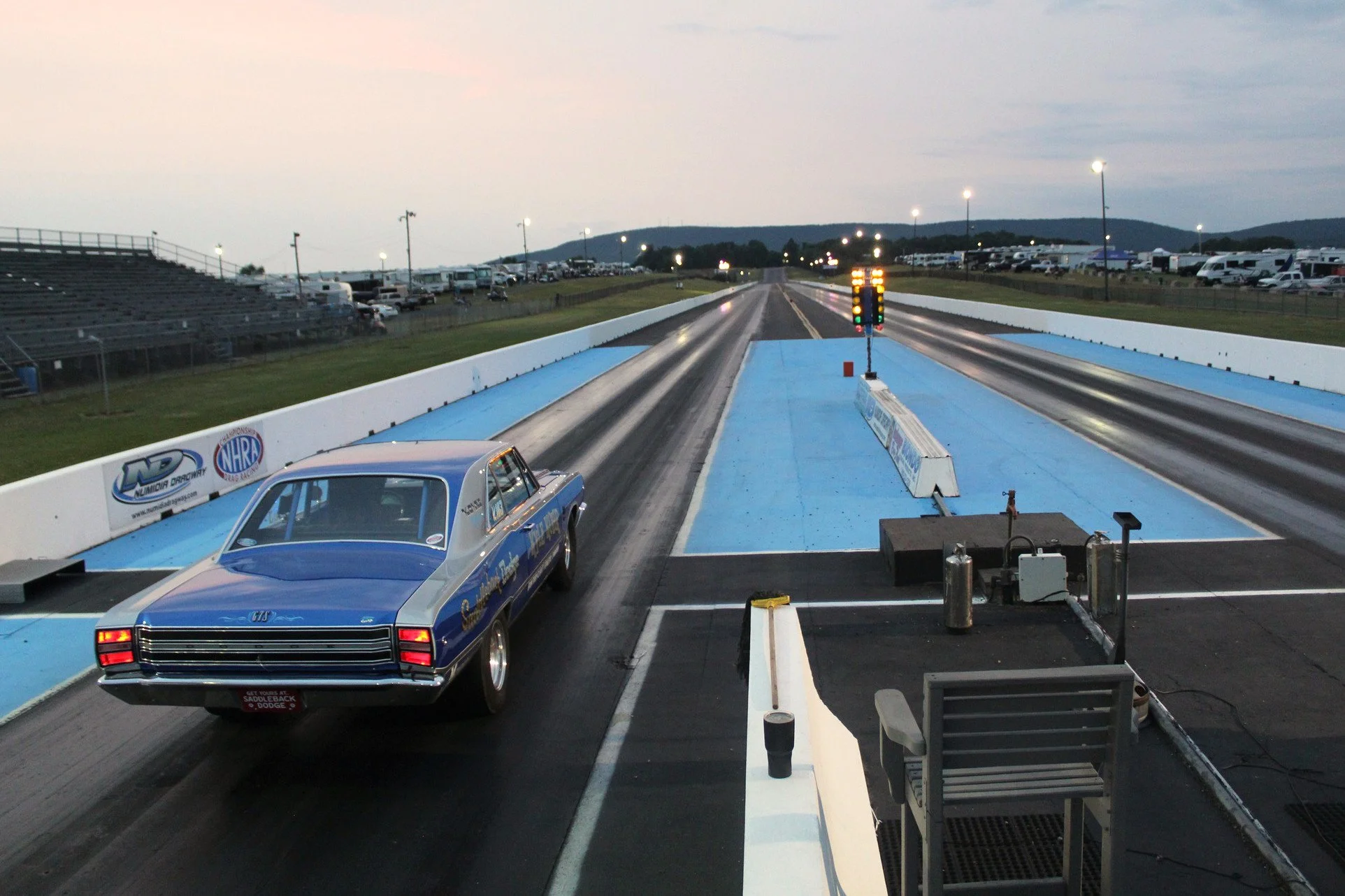 A drag racing track with a blue and white race car on the starting line and a finish line with a light tree, surrounded by parking lots and hills in the background, during dusk.