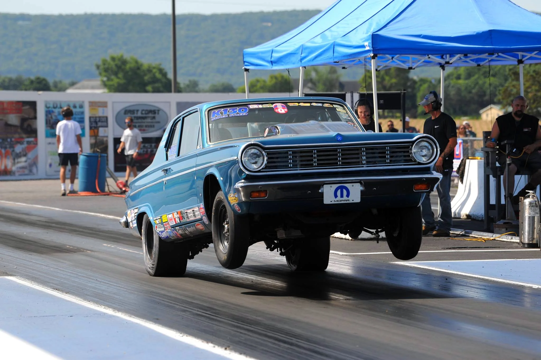 A blue vintage drag racing car with sponsor stickers is performing a wheelie on a drag strip during a race, with crew members and spectators in the background under a blue canopy.