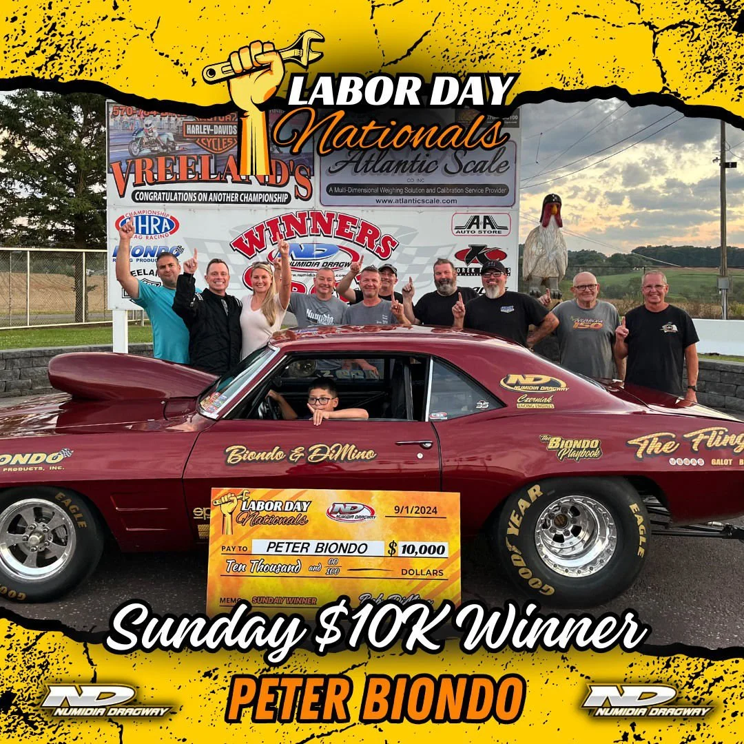 A group of people celebrating as a young boy, Peter Biondo, sits in a racing car holding a large check for $10,000 for winning the Sunday Labor Day Nationals at Numidia Dragway. The group is standing behind the car, raising their hands in victory, an