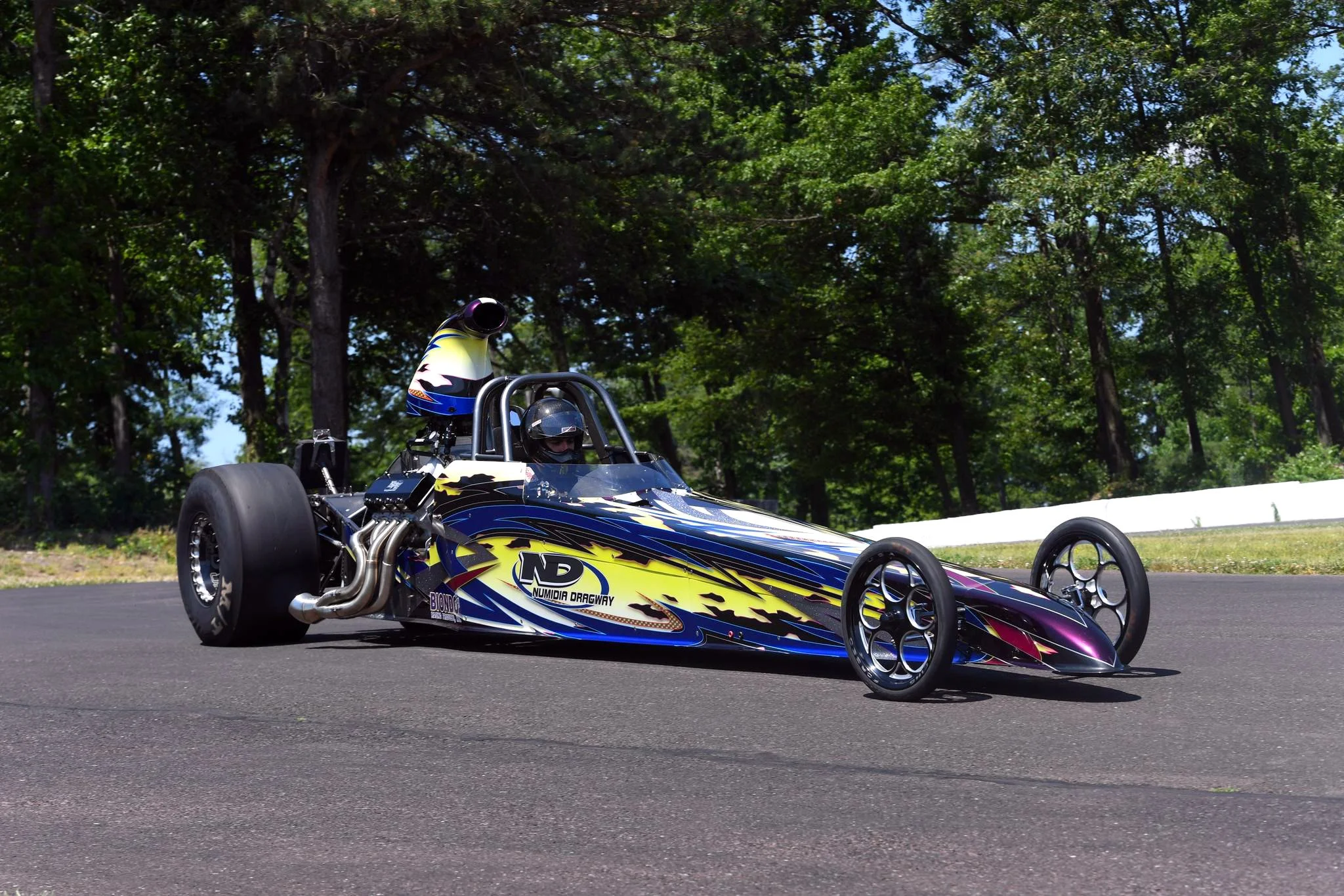 A single-seat race car with a colorful yellow, purple, and blue paint job, featuring the words "Numidia Dragway" on the side, is on a paved track surrounded by green trees, with a driver wearing a helmet seated inside.