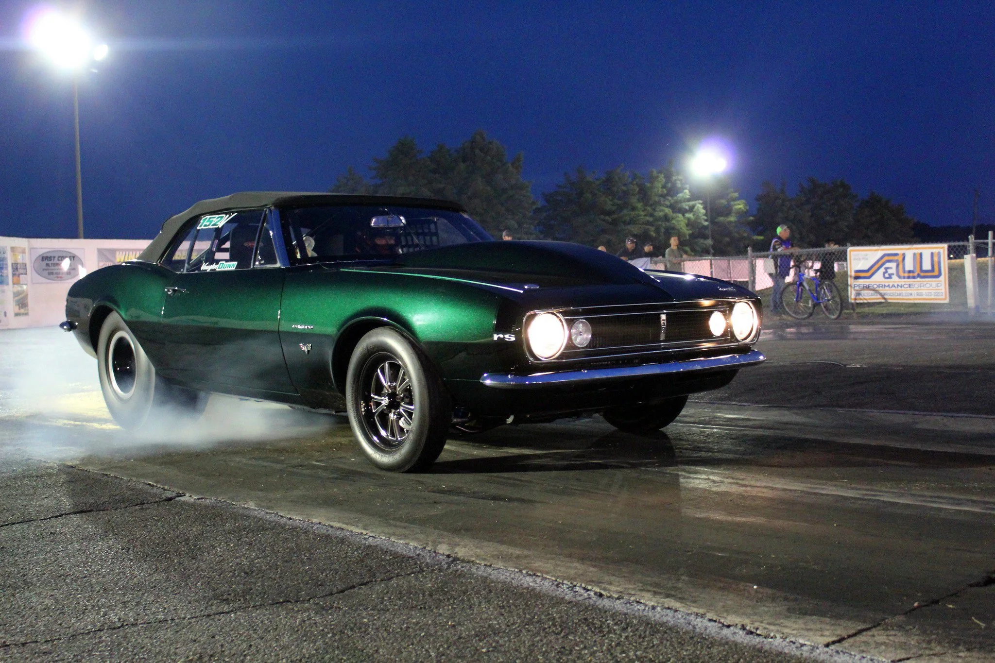 A green vintage muscle car performing a burnout at night, with tire smoke and bright stadium lights in the background.