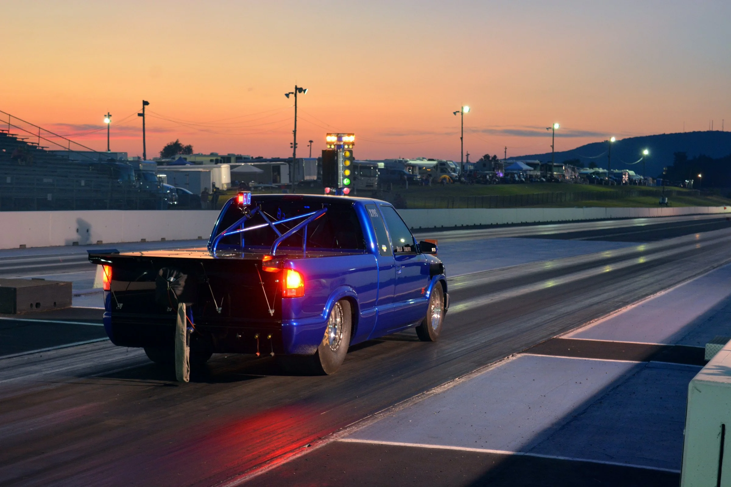 A race car on a drag strip during sunset with a lighted Christmas tree-style Christmas tree starting tree in the background and spectators and trailers in the distance.