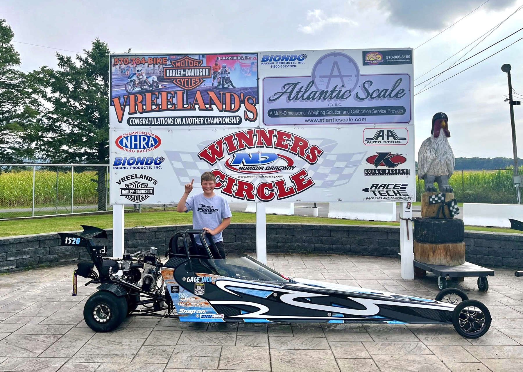 A young boy standing next to a drag racing car at a race track. The boy is smiling and making a peace sign with his right hand, wearing a gray t-shirt. The background includes a large sign with multiple advertisements for motorcycle shops and racing supplies, a statue of a bird on a wooden stand, and a green field with trees.