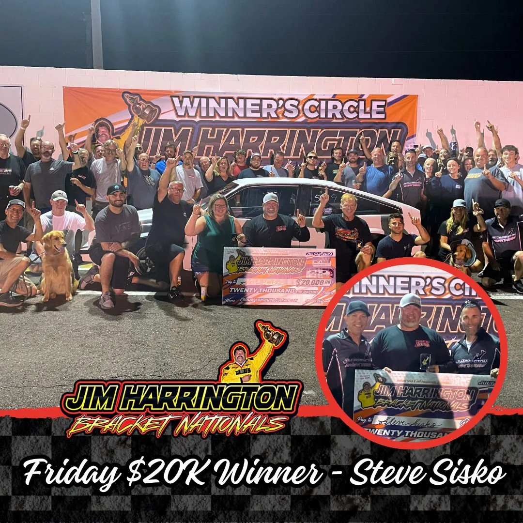 A large group of people gathered outdoors at night, celebrating a victory with a race car in the center. The group holds up their hands, some holding drinks, and they are smiling. A banner behind them reads 'Winner's Circle Jim Harrington'. In the fo