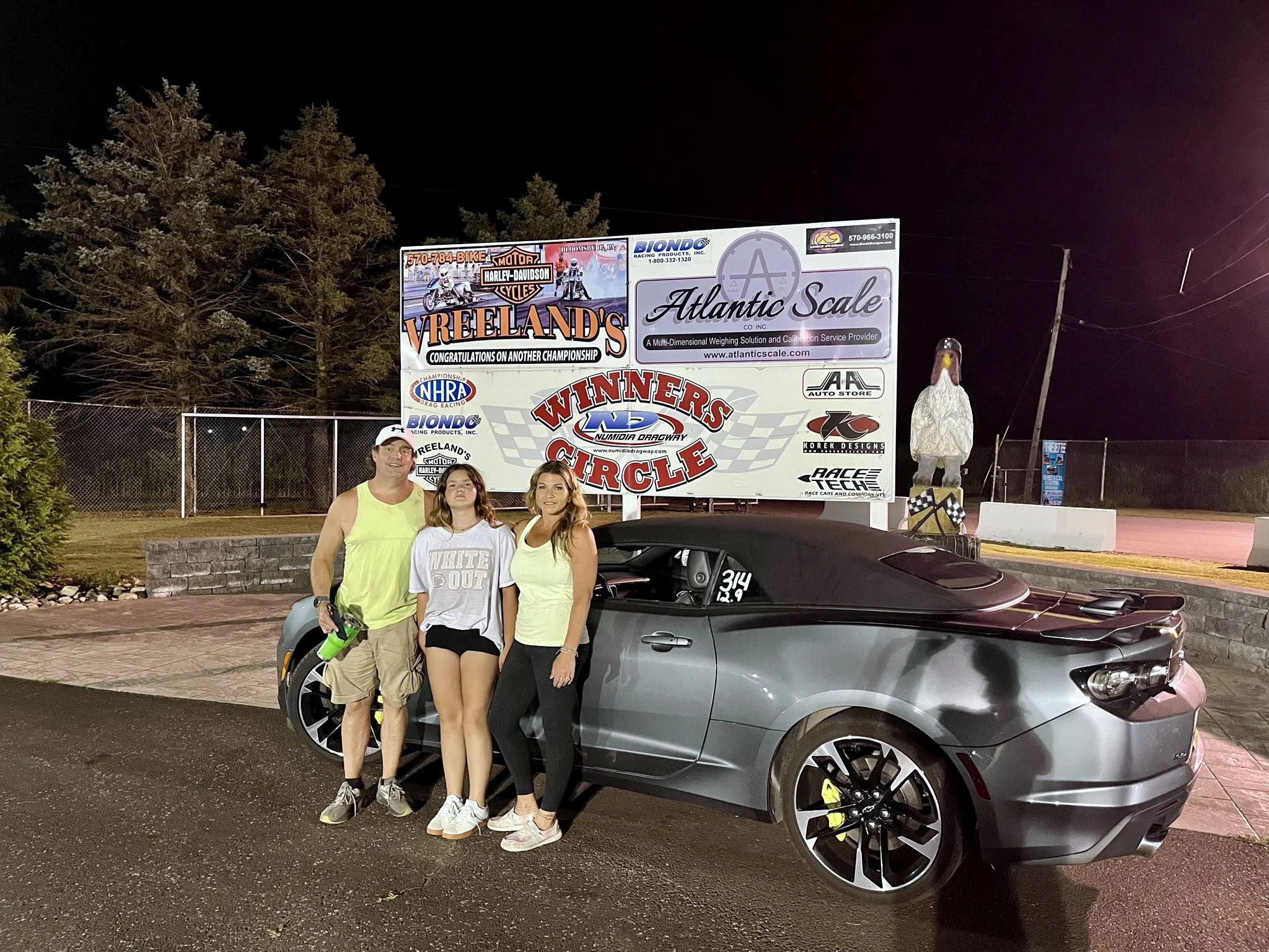 A group of three people, two women and one man, standing next to a gray sports car at night. They are in front of a large billboard advertising drag racing and motorcycle events, with trees and utility poles in the background.
