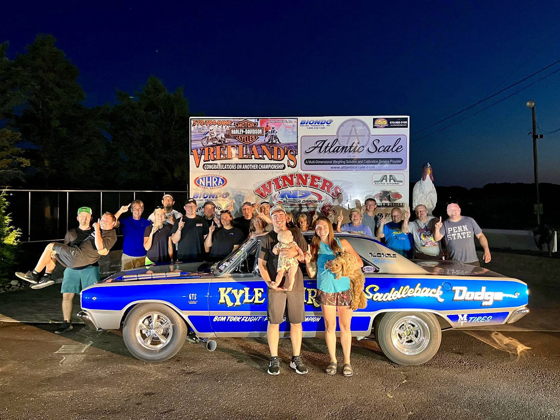 Group of people standing beside a blue drag racing car in front of a large sponsor banner at night. The group is smiling and making celebratory gestures.