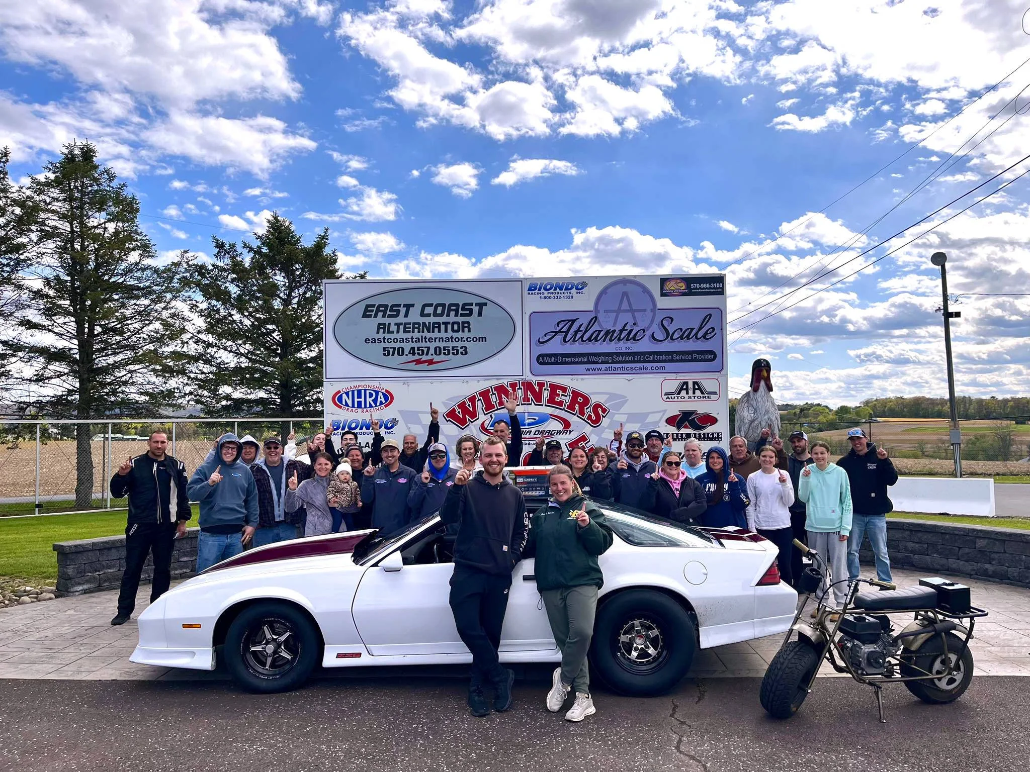 Group of people gathered outdoors at a racing event, posing in front of a racing sign and a white racing car, with a clear cloudy sky and trees in the background.