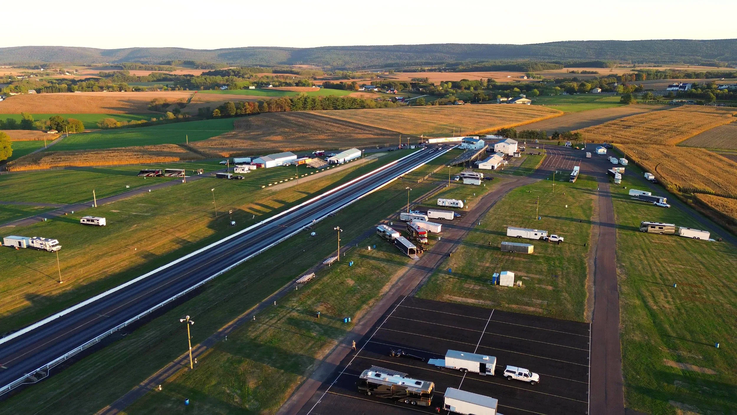 An aerial view of a drag racing strip surrounded by grassy fields and parked RVs and trailers, with a rural landscape and hills in the background.
