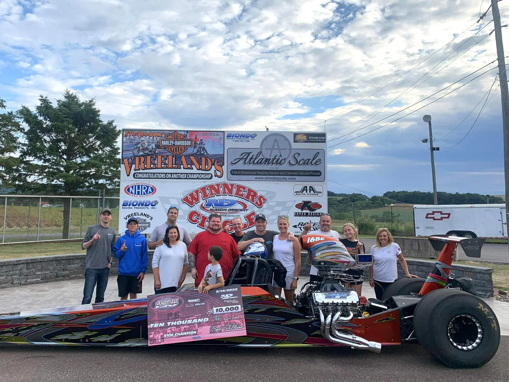 A group of people standing next to a race car at a racing event. There are signs and banners in the background, including a large Vreeland's banner, a sponsor Atlantic Scale, and others. The race car has a checkered flag design and a large prize chec