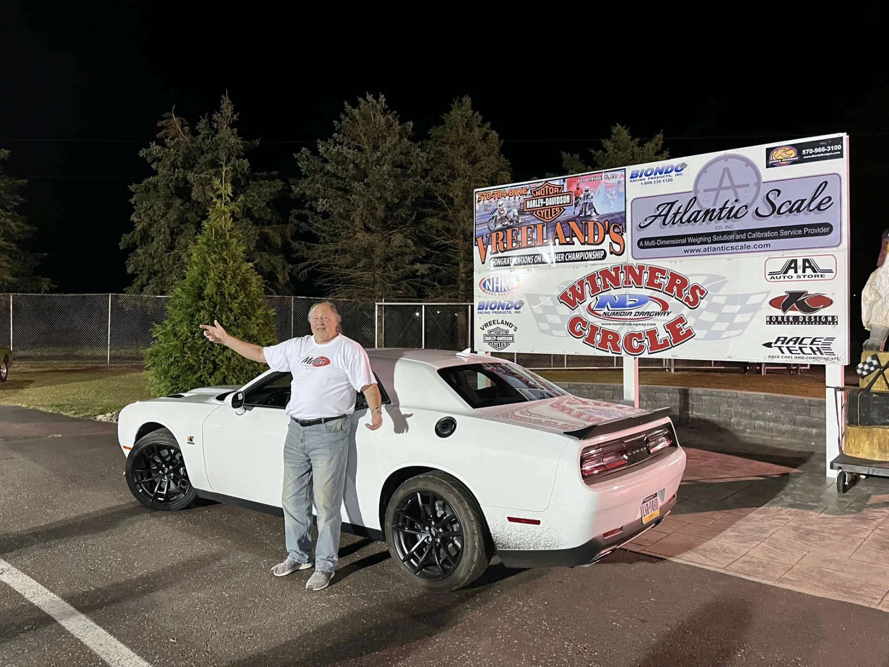 A man with gray hair and glasses wearing a white t-shirt and gray pants stands next to a white sports car with black rims at night. Behind him, there is a large sign for Vreeland's Motor Harley-Davidson Cycles, Atlantic Scale, and other sponsors, and