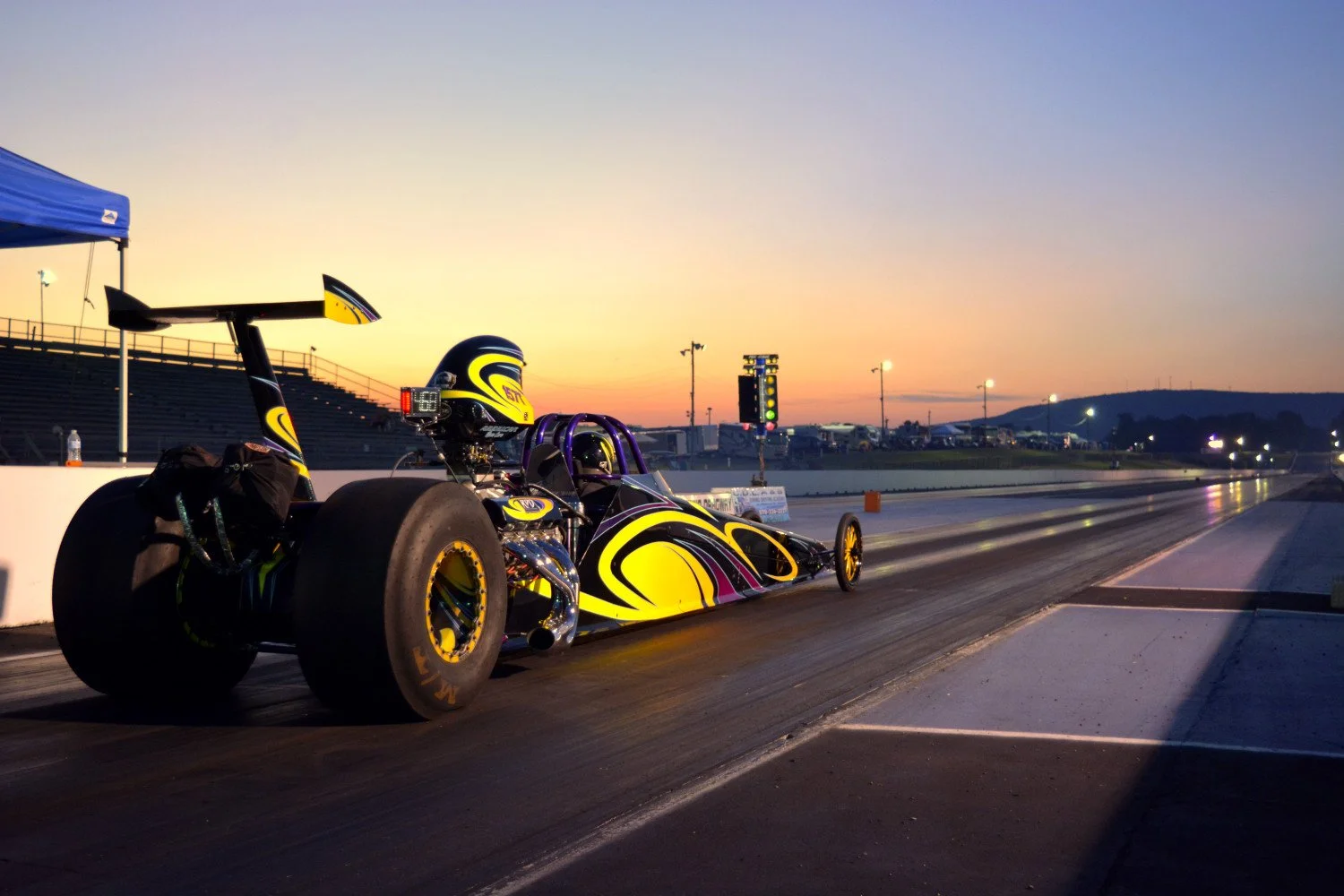 Race car on a drag strip during sunset with empty bleachers, a tent, and a race scoreboard in the background.