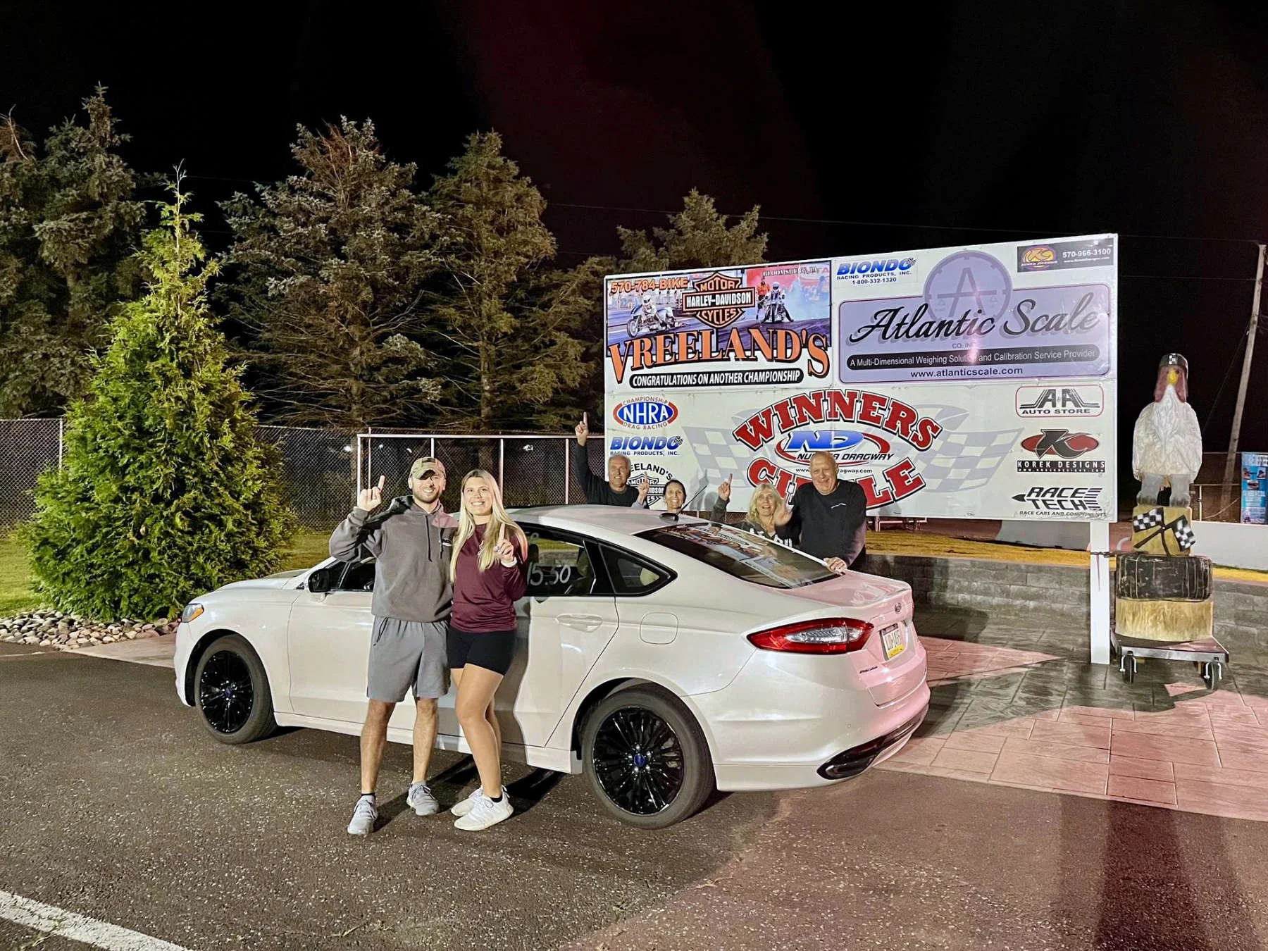 Group of five people standing next to a white sports car at night, posing with their middle fingers raised, in front of a racing scoreboard, sign, and inflatable turkey decoration at a racing event.