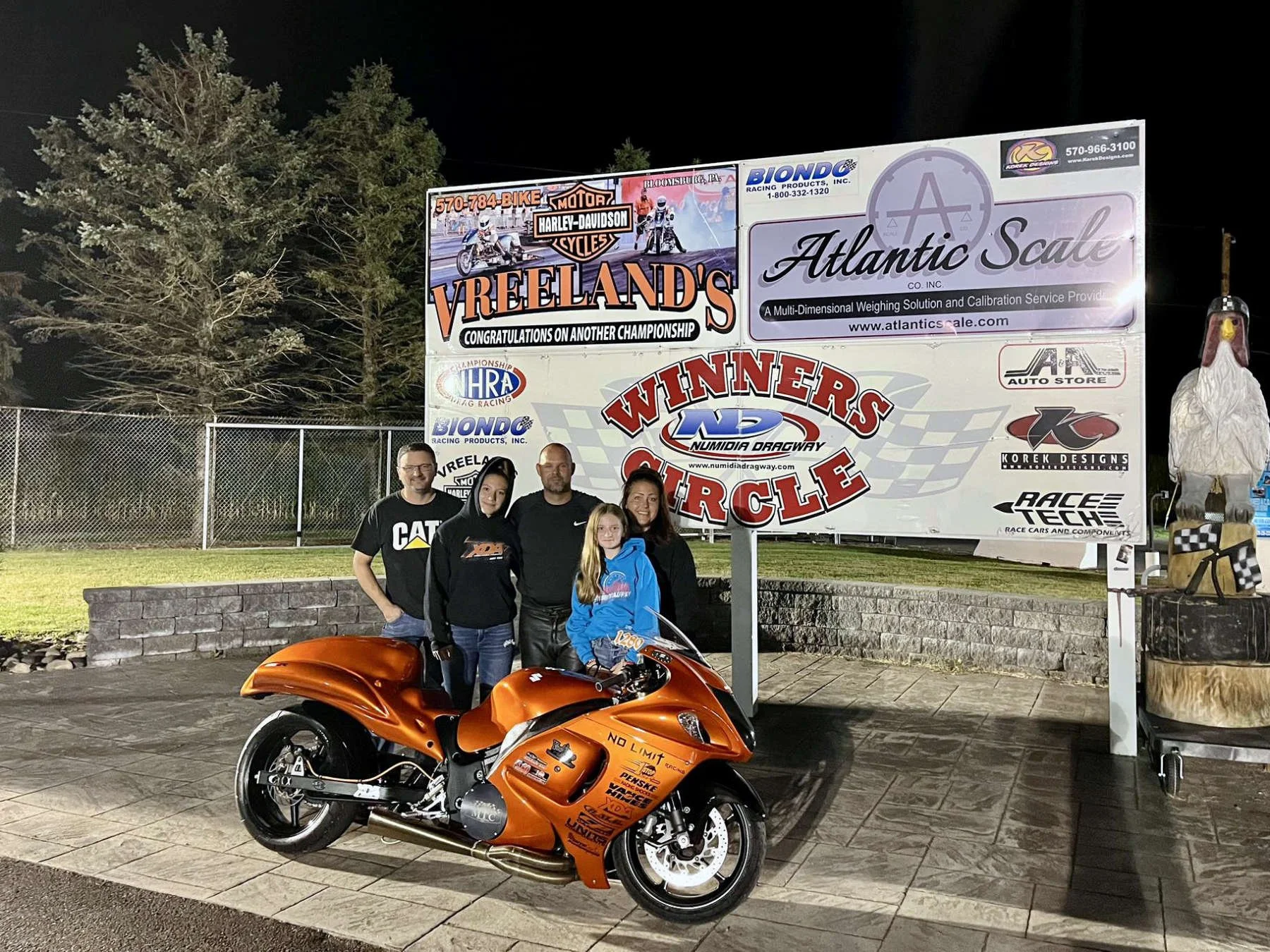 A group of five people standing outdoors at night, posing in front of a large sign and an orange sports motorcycle. The sign has various advertisements for local businesses including Vreeland's, Atlantic Scale, and others, along with racing-related l