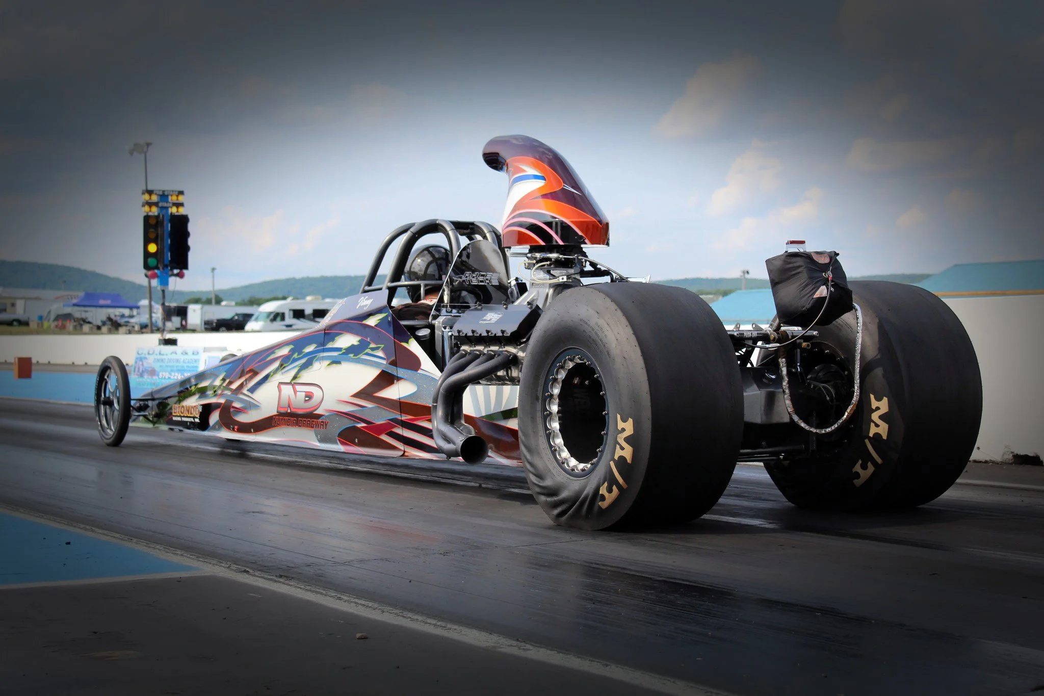 Image of a drag racing car on the track with large rear tires and a painted aerodynamic helmet in front of a cloudy sky.