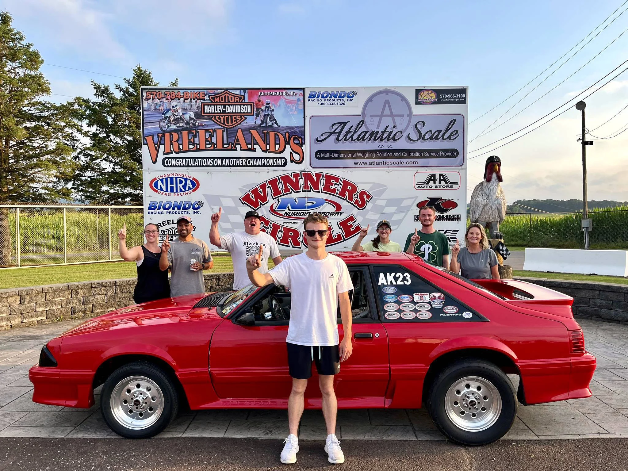 Group of people standing in front of a red car and a large billboard at a racing event. The billboard features Harley-Davidson and Atlantic Scale advertisements, and the group is smiling and posing for the camera.