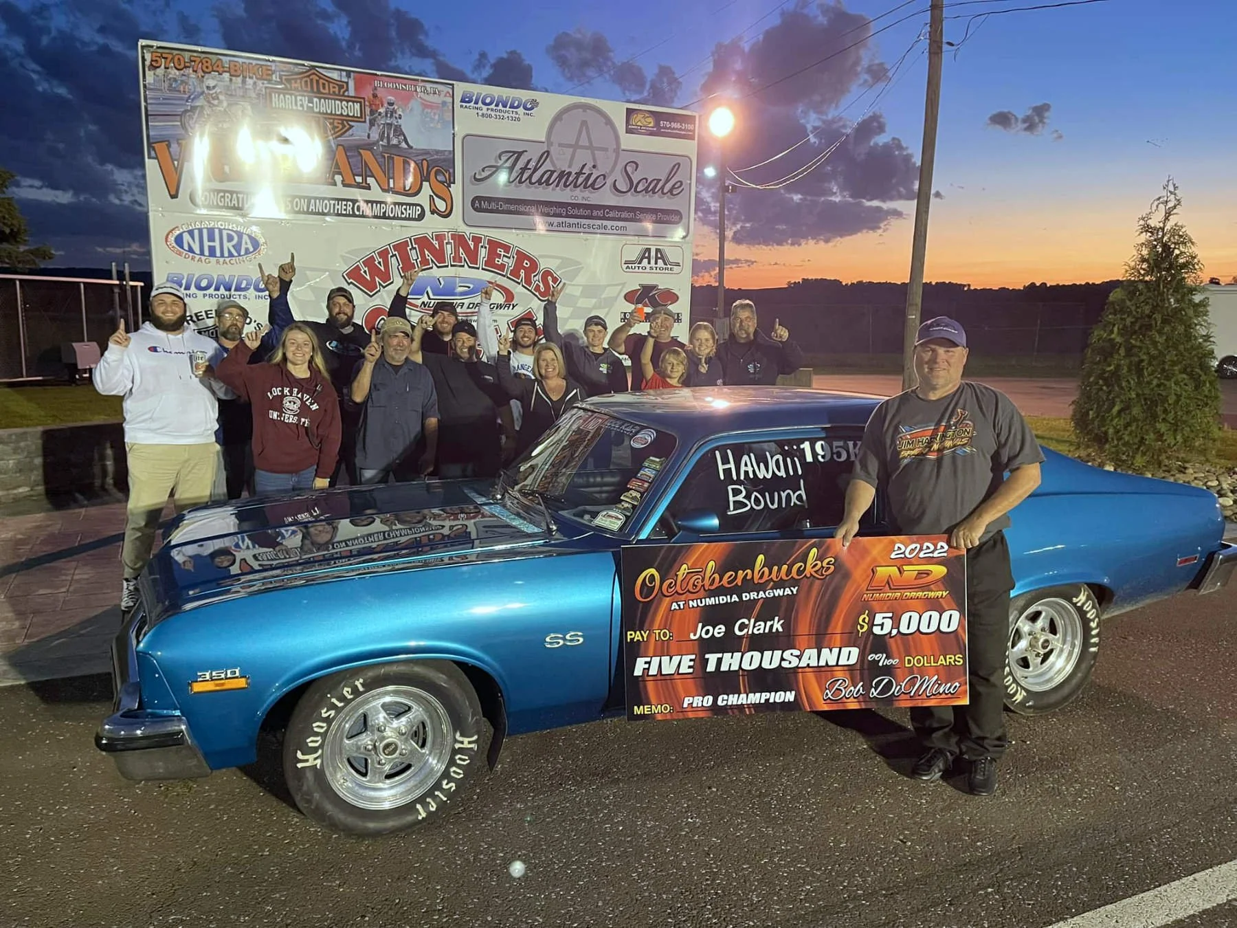 A group of people celebrating around a blue vintage car at dusk, holding a large check for $5,000. The car has a sign that says 'Hawaii Bound' and the background features a large sign with various sponsors and a sunset sky.