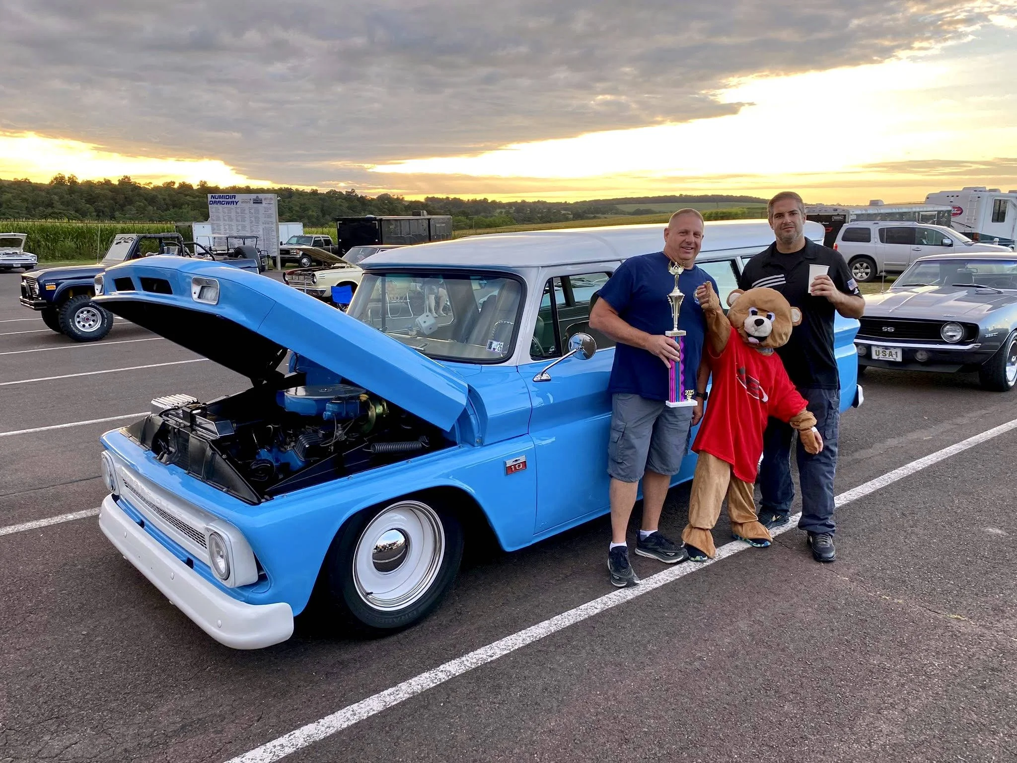 Two men and a young person dressed as a bear, standing next to a blue vintage drag racing car, with a trophy and drink, at a car show during sunset.