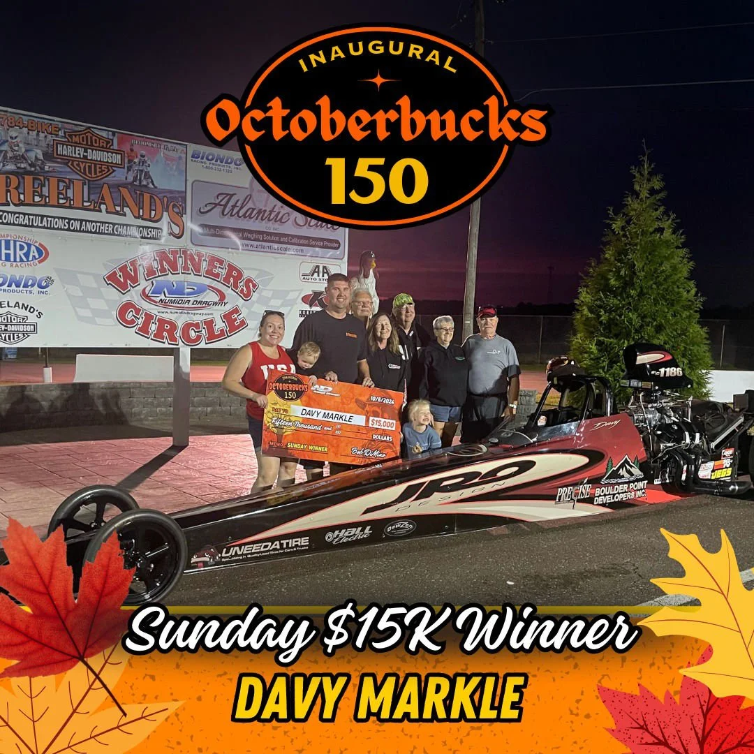 A group of people holding a large $15,000 check at a race track shows a racing team celebrating their win. The check is made out to Davy Markle, and a racing car is in the foreground. The backdrop features banners and a sign celebrating the inaugural