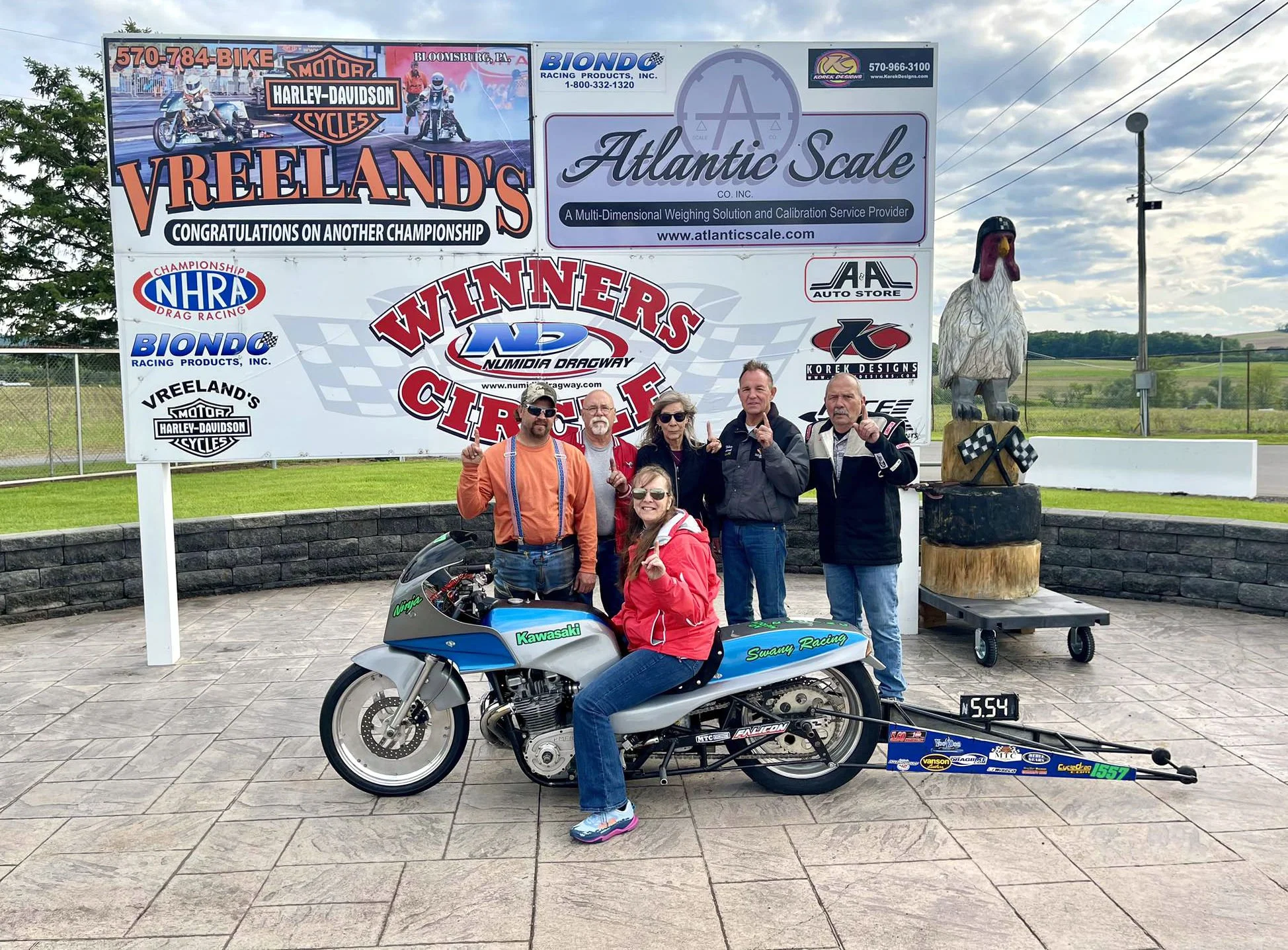 A group of people standing outdoors in front of a large sign with various advertisements and sponsors, including Atlantic Scale and Vreeland's. Two women are sitting on a race motorcycle with a girl in a red jacket sitting nearby. One of the women is wearing glasses and a red jacket, while the other woman has long curly hair and sunglasses. Four men are standing behind them, some pointing upwards. There is a large chicken sculpture on a wooden stand to the right of the sign. The background shows a grass field and cloudy sky.