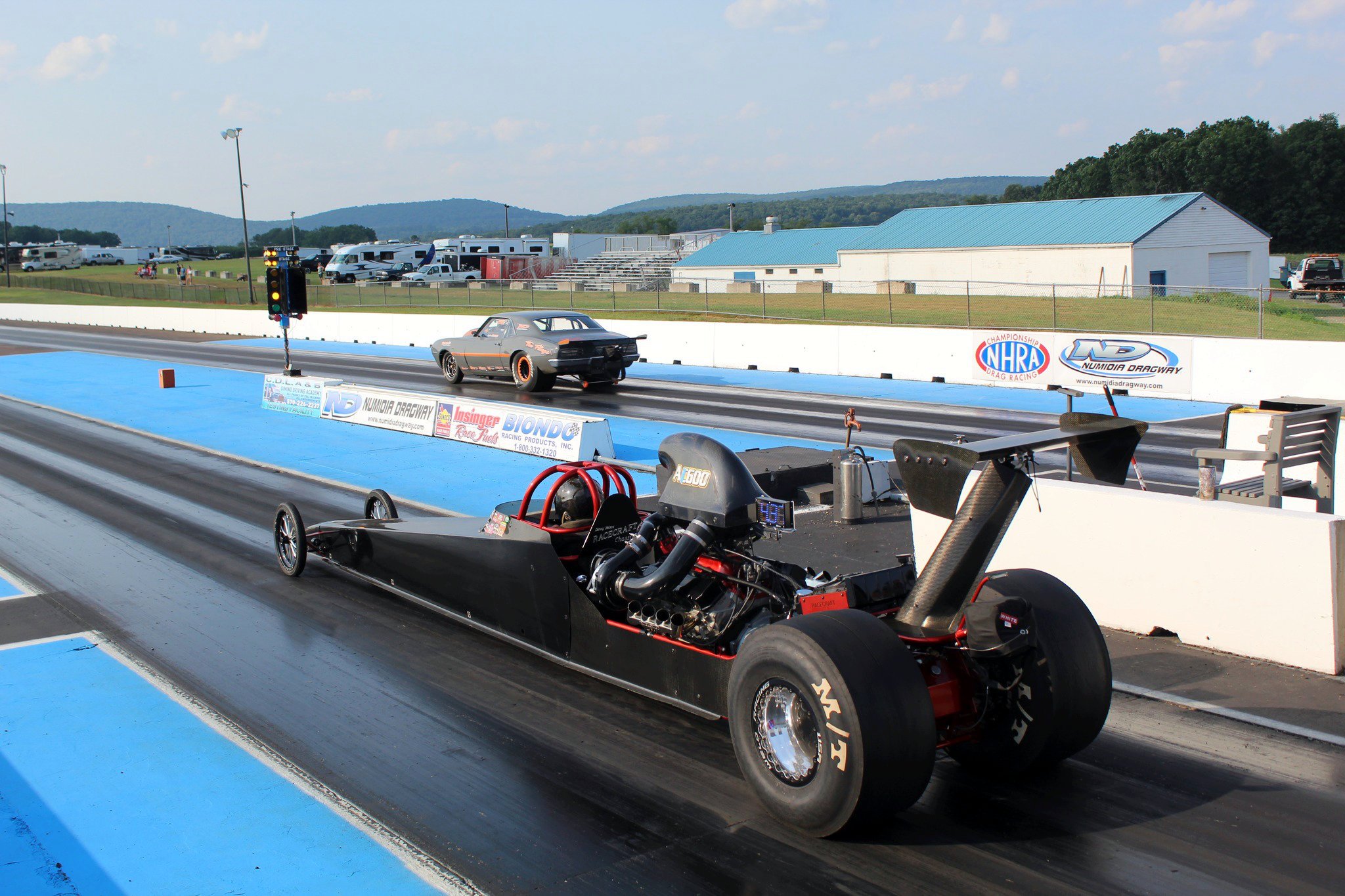 A drag racing car on the starting line at Numidia Dragway with equipment and vehicles in the background, including a fight track and trailers.