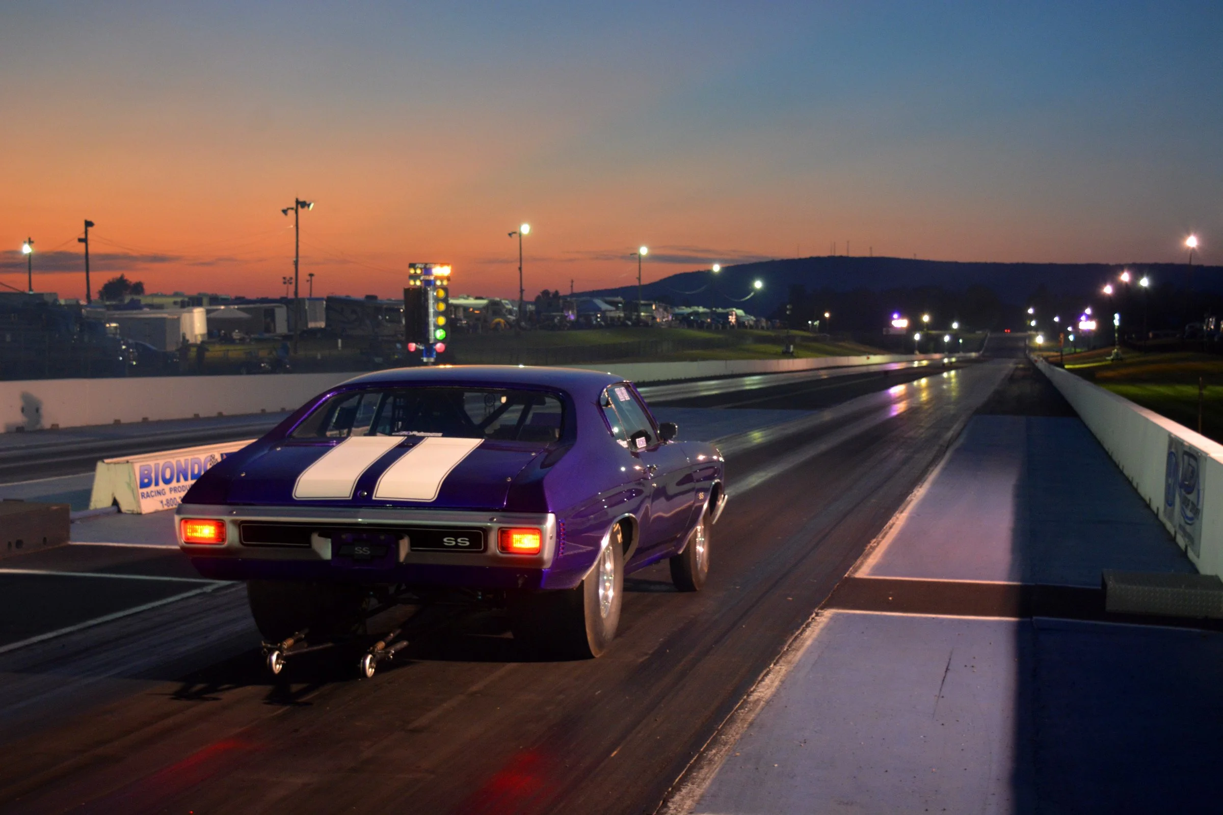 A purple race car with white stripes on a drag strip at dusk, with bright lights illuminating the track and a row of trailers and tents in the background.