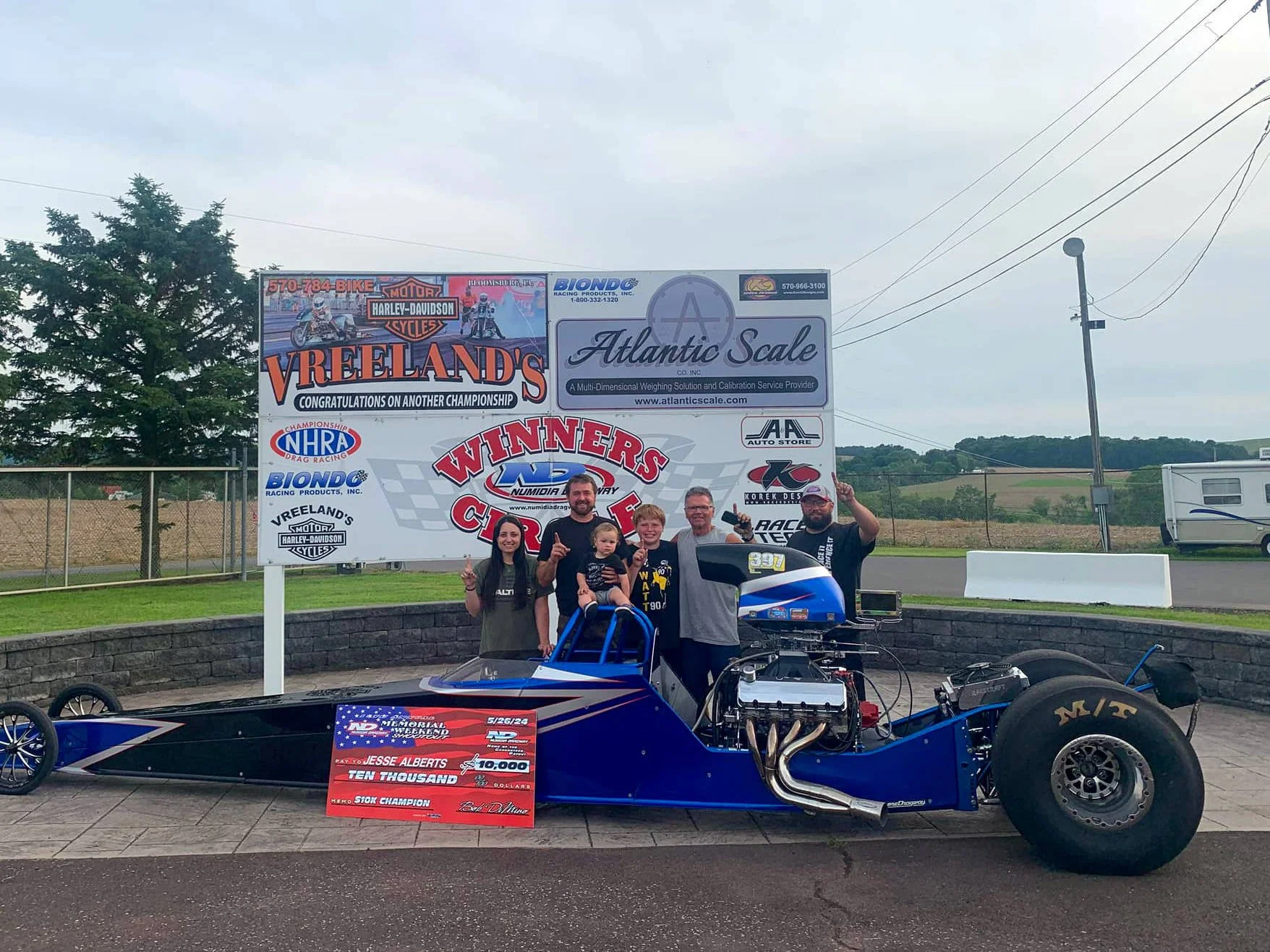 A group of six people, including children and adults, posing next to a racing car at a motorsports event. The background features a large signboard with various sponsor logos and congratulatory messages, with a race track and trailers in the distance