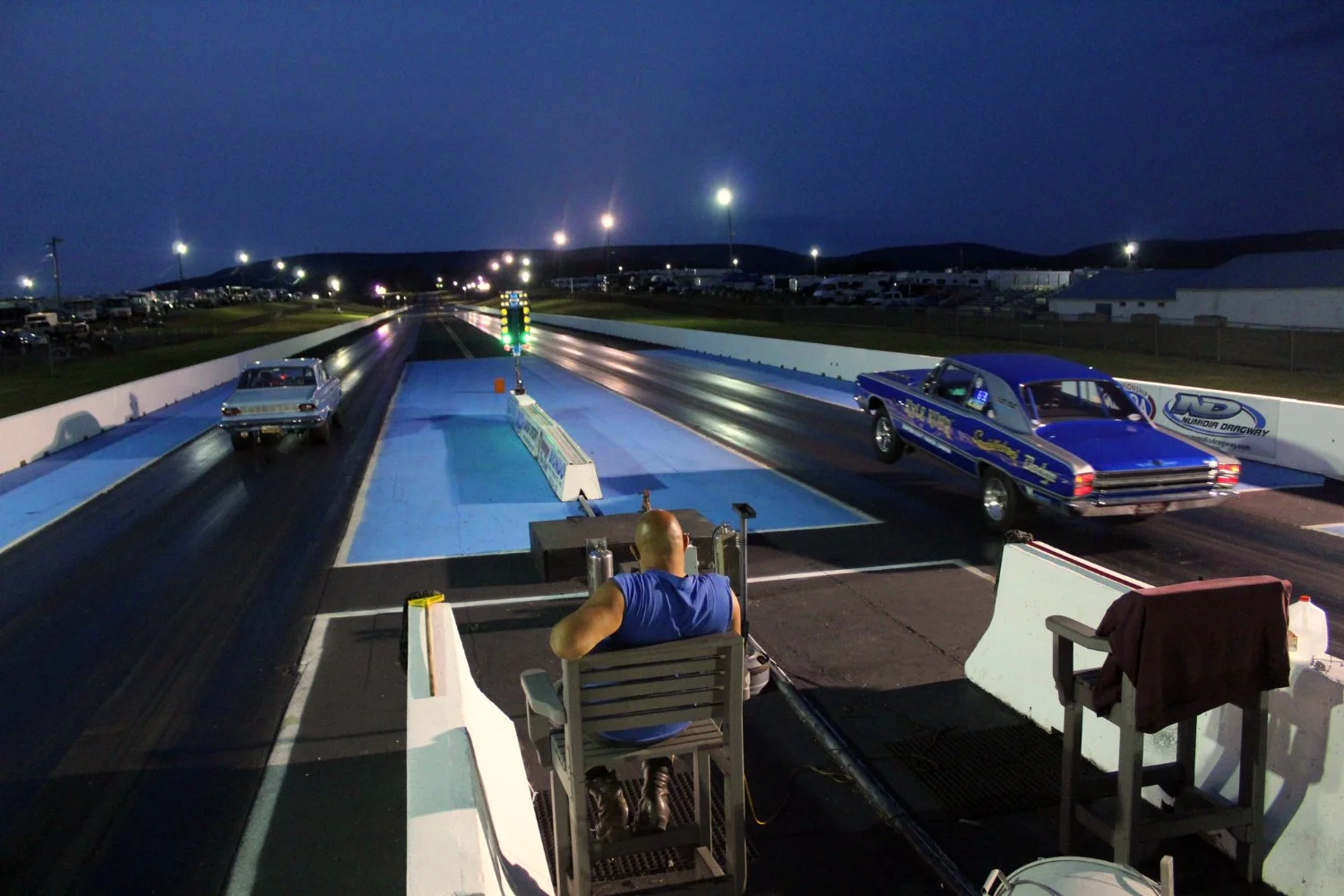 Nighttime shot of a drag racing strip with two cars, one in the right lane and the other on the left, preparing for a race, with a man seated observing from a chair in the foreground.