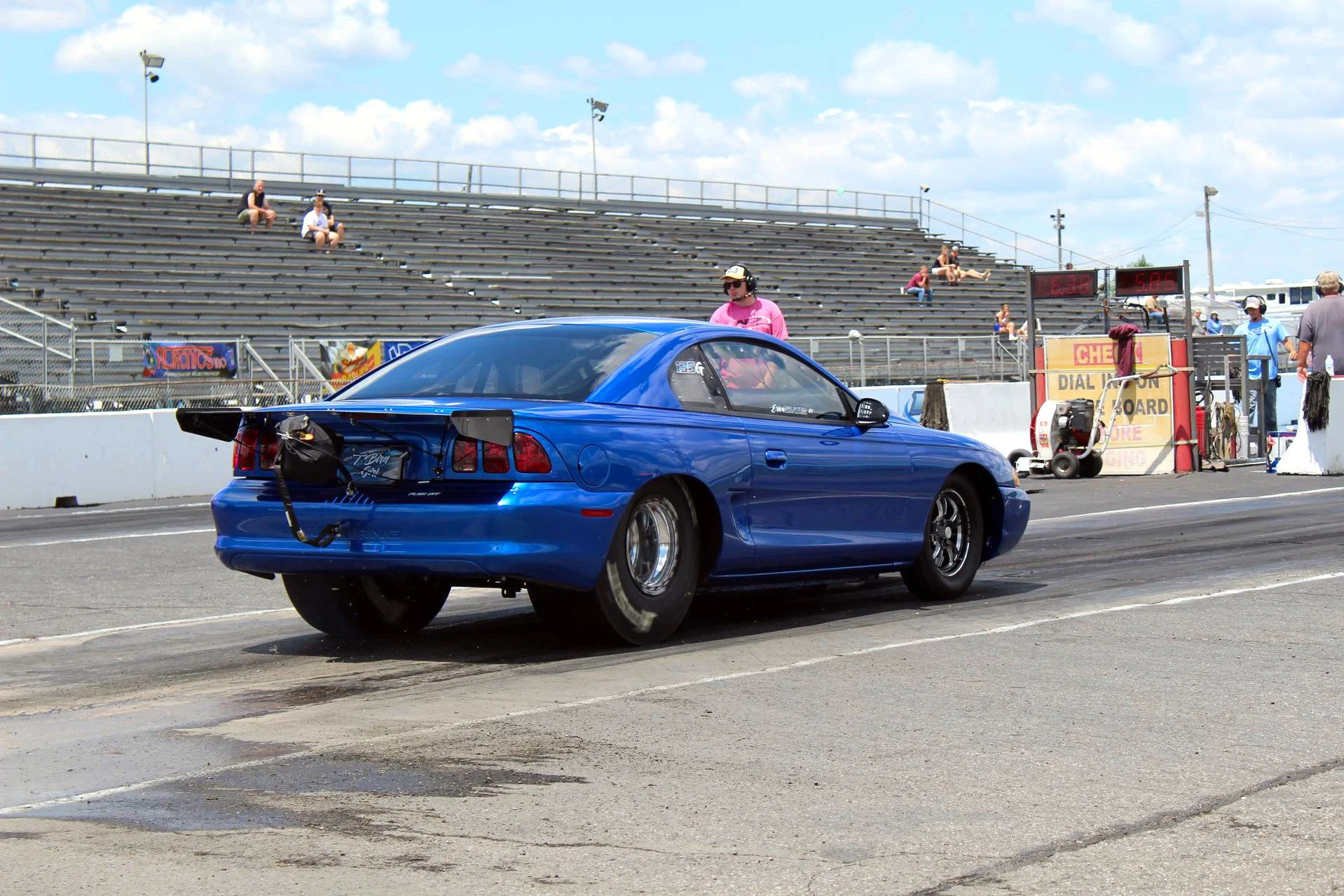 Blue sports car at a drag racing strip during daytime, with spectators sitting on bleachers and crew members near the finish line.