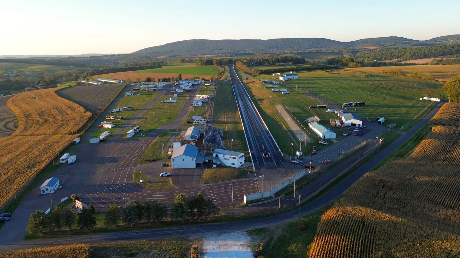 Aerial view of a rural area with a drag strip, multiple RVs and trailers, farm fields, and hills in the background during sunset.