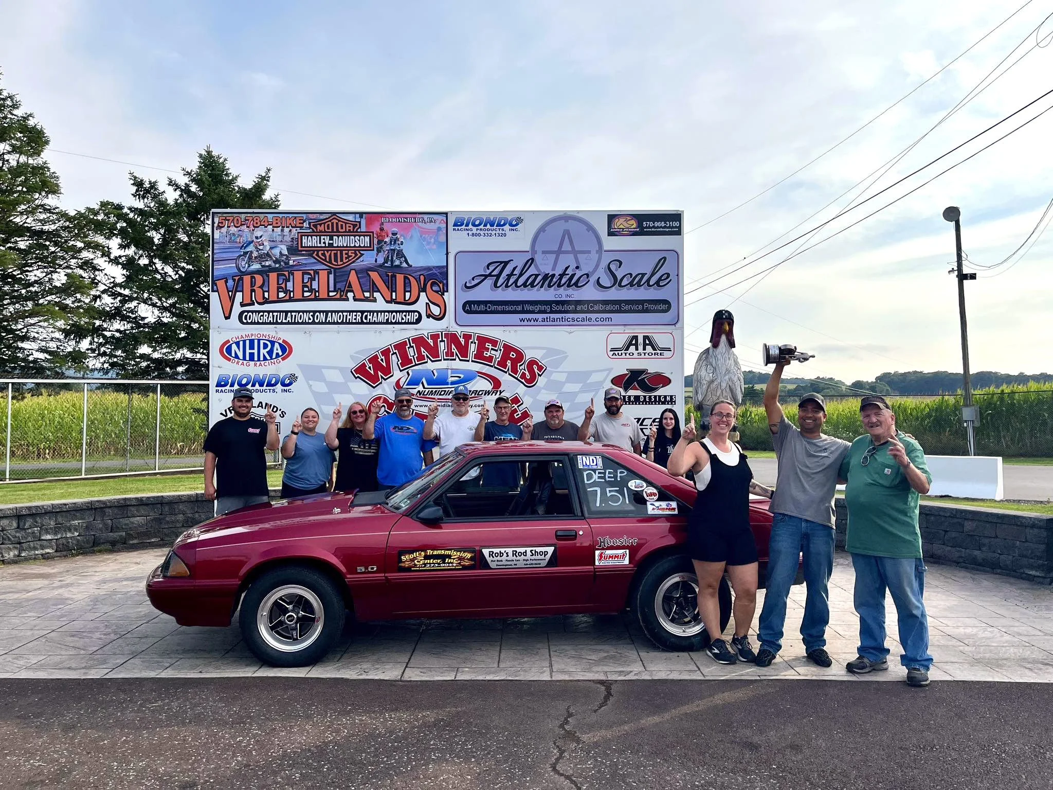 Group of ten people standing in front of a racing car and a large winner circle with advertisements and sponsors, outdoors on a paved area with trees and power lines in the background.