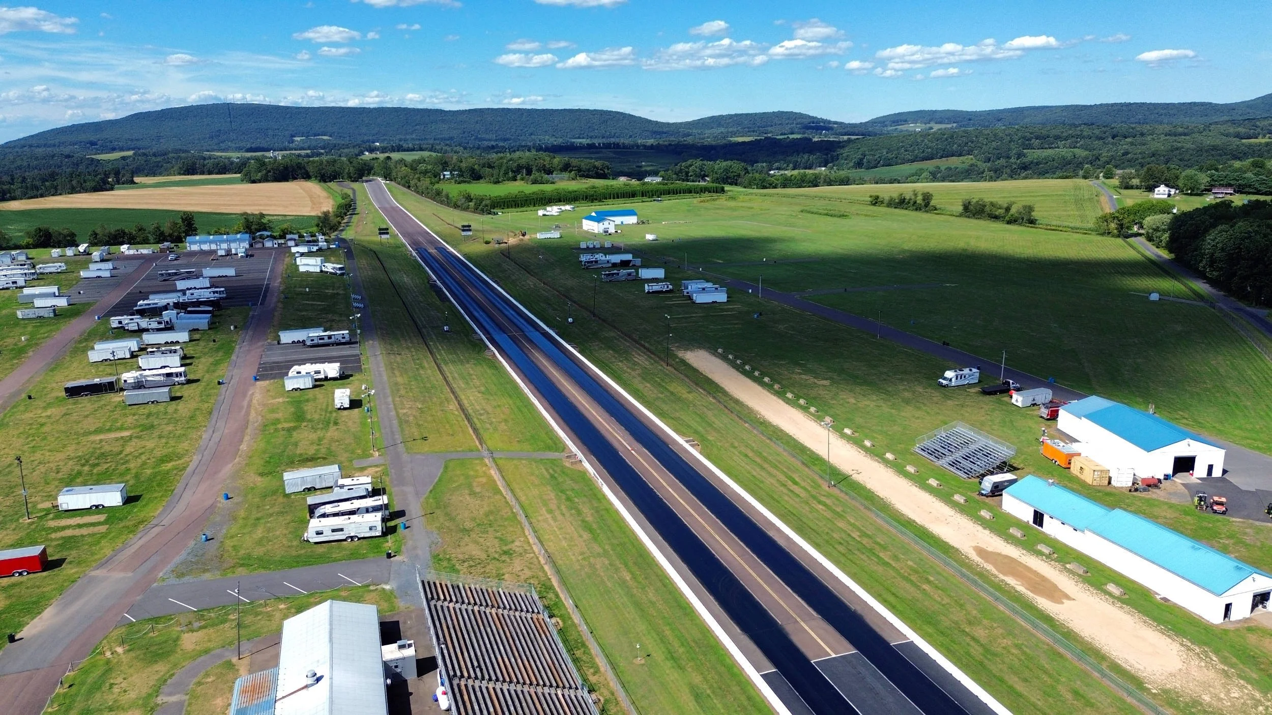 Aerial view of a rural area with fields, roads, and parked trailers and vehicles, with mountains in the background under a partly cloudy sky.
