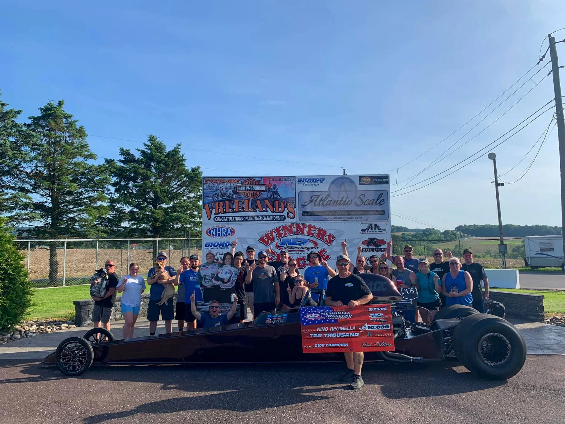 A large group of people posing for a photo outdoors, with a race car in the foreground and a sign in the background celebrating winners of a racing event. The scene is set on a sunny day with trees and hills in the distance.