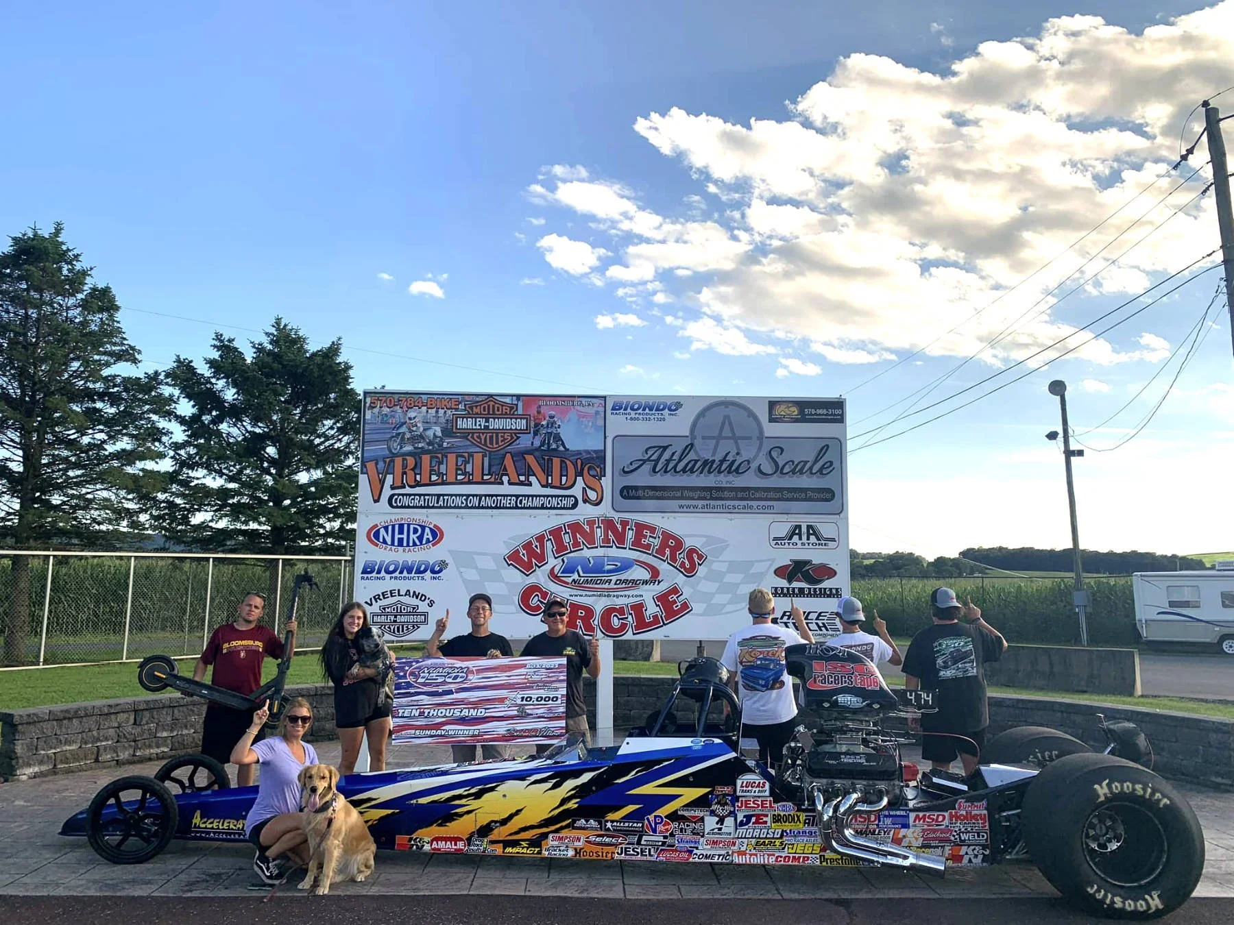 People at a drag racing event holding a large check and posing next to a dragster car with various sponsor decals. A sign with event details and sponsors is visible in the background, along with trees, power lines, and a blue sky with scattered cloud