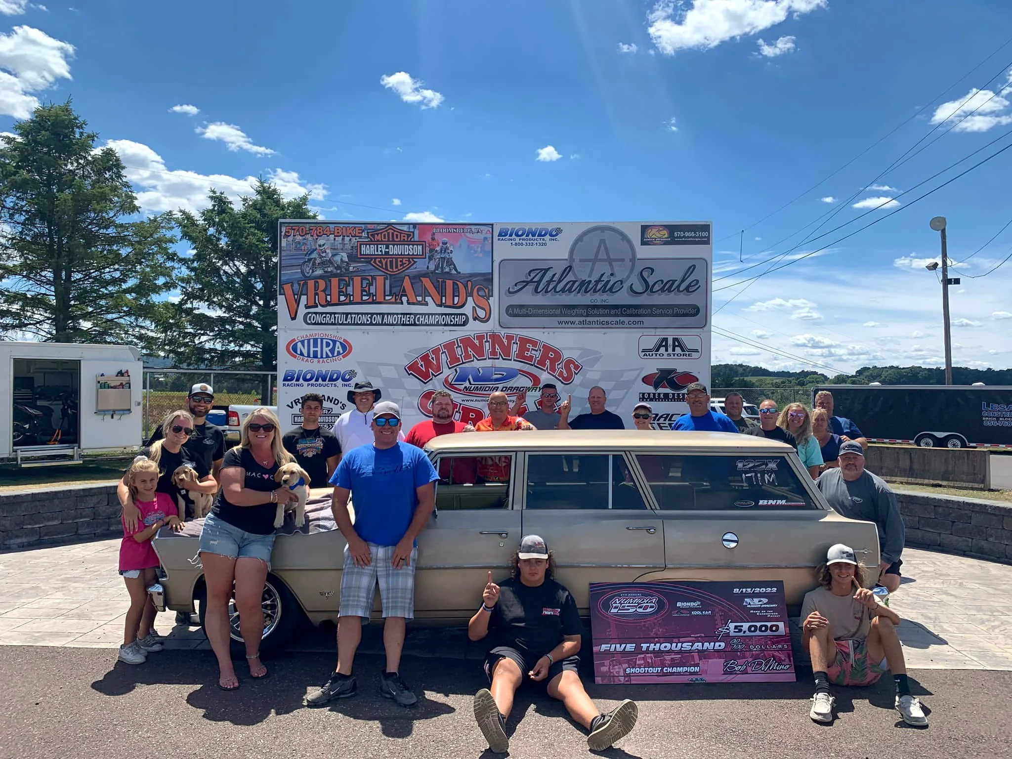 A group of people posing in front of a classic car with a large checkboard for a racing event, with banners and trees in the background, under a partly cloudy sky.