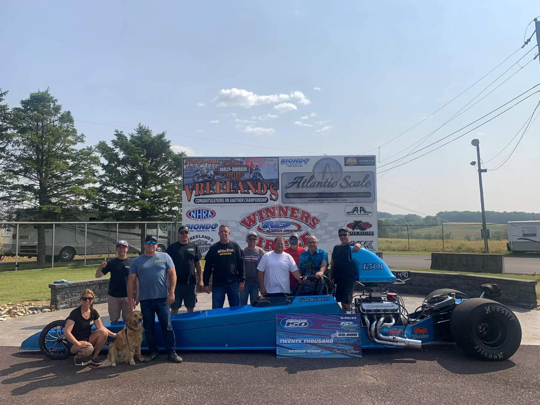 Group of people posing next to a blue drag racing car at a race track, with banners and trees in the background.