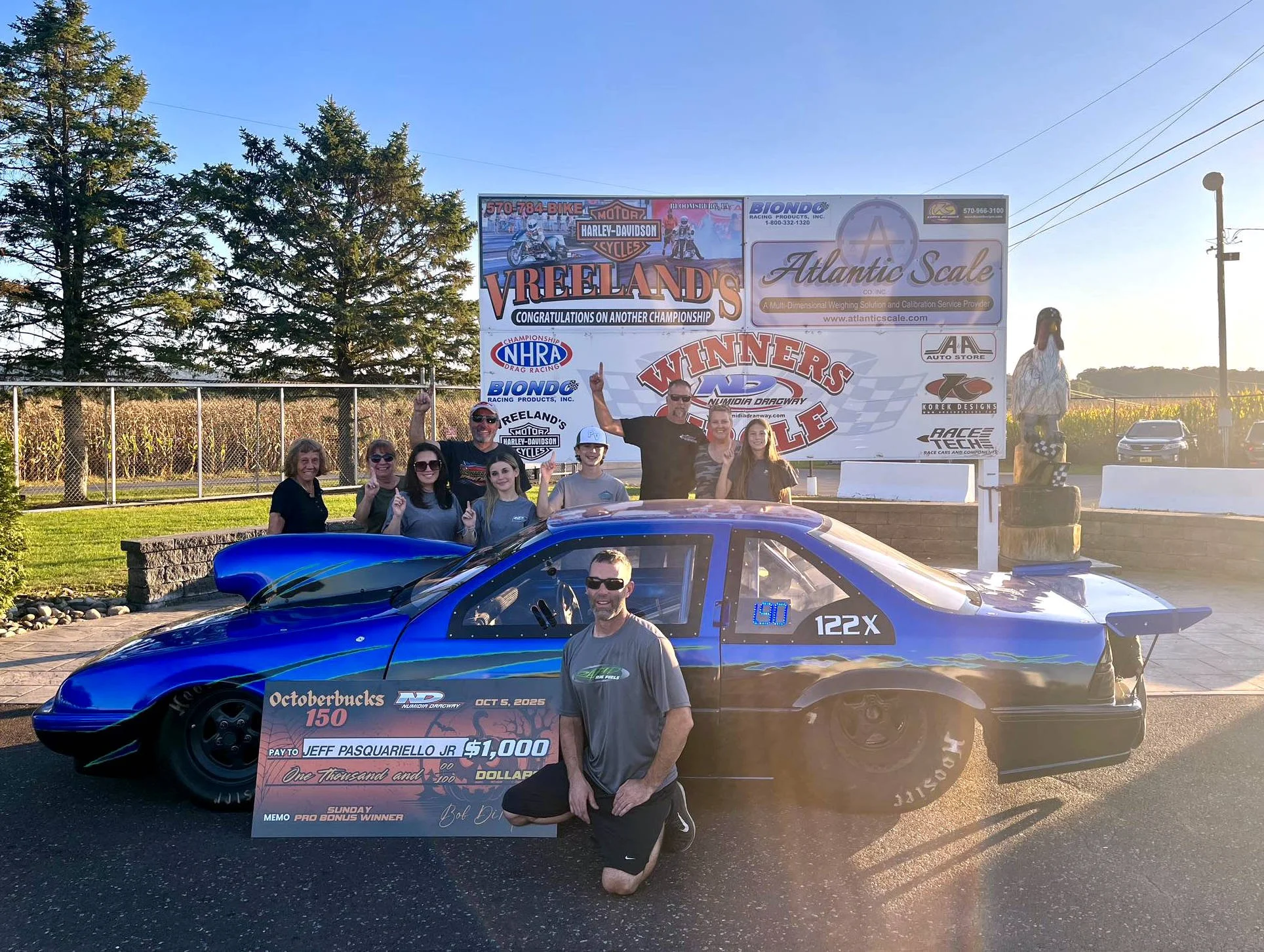 Group of people celebrating at a racing event next to a blue race car holding a large check for $1,000. The background features banners with racing and sponsorship logos, including Harley-Davidson, NHRA, Atlantic Scale, and others.