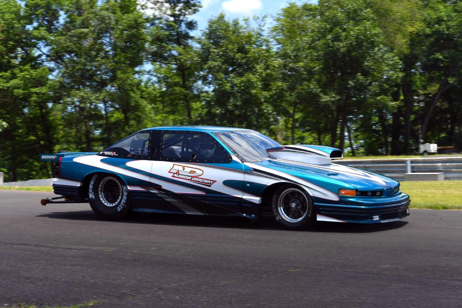 A blue, black, and white race car on a track with green trees in the background.