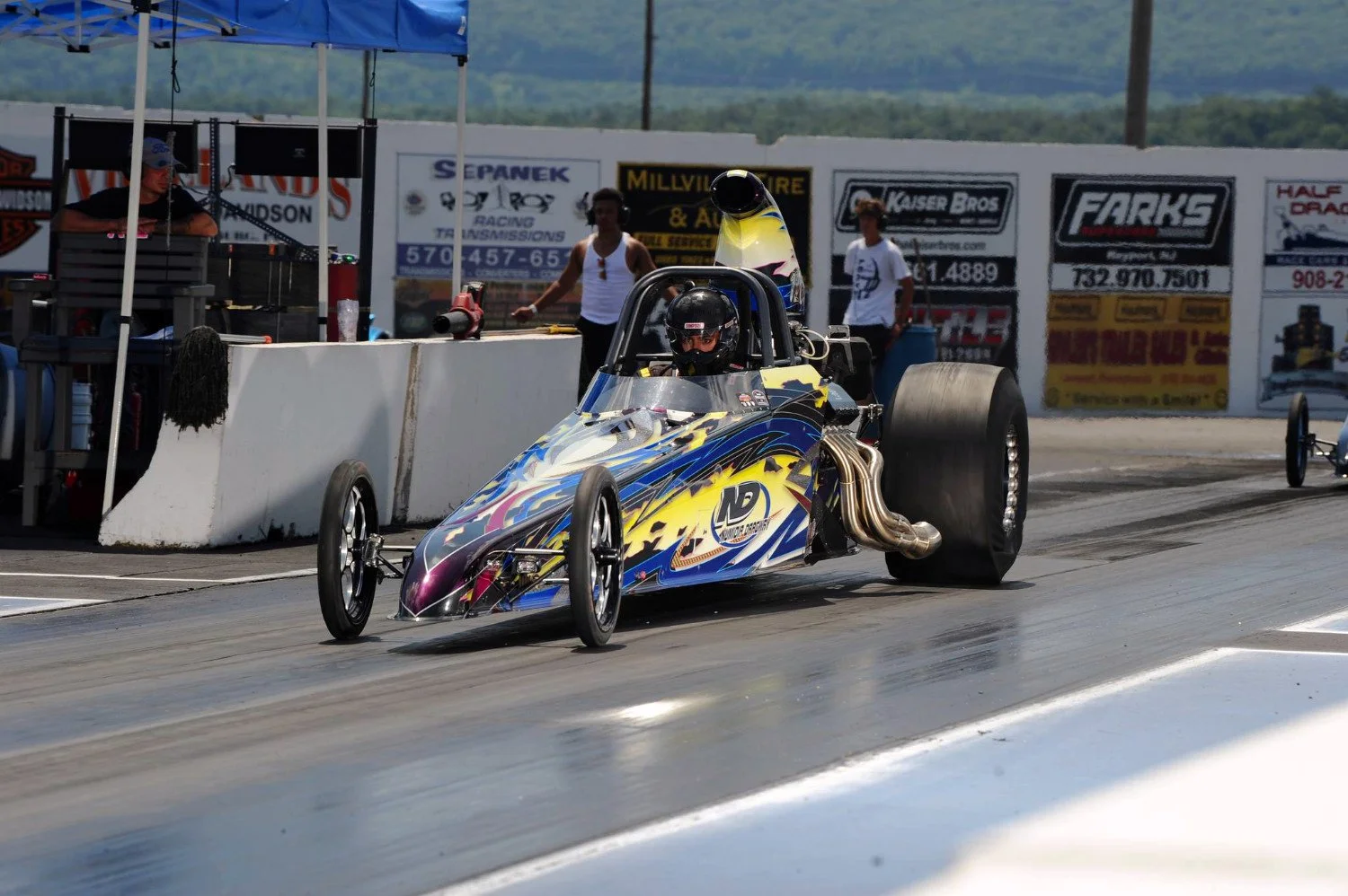 Drag racing car on the starting line with a driver wearing a helmet, surrounded by people and advertisements in the background.