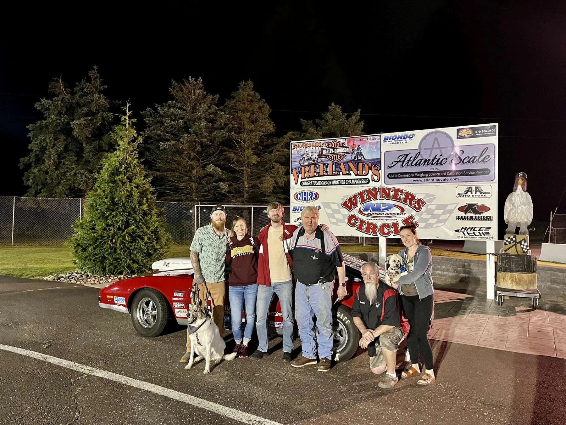 Group of six people and two dogs standing in front of a red race car at night, with a large billboard and trees in the background.