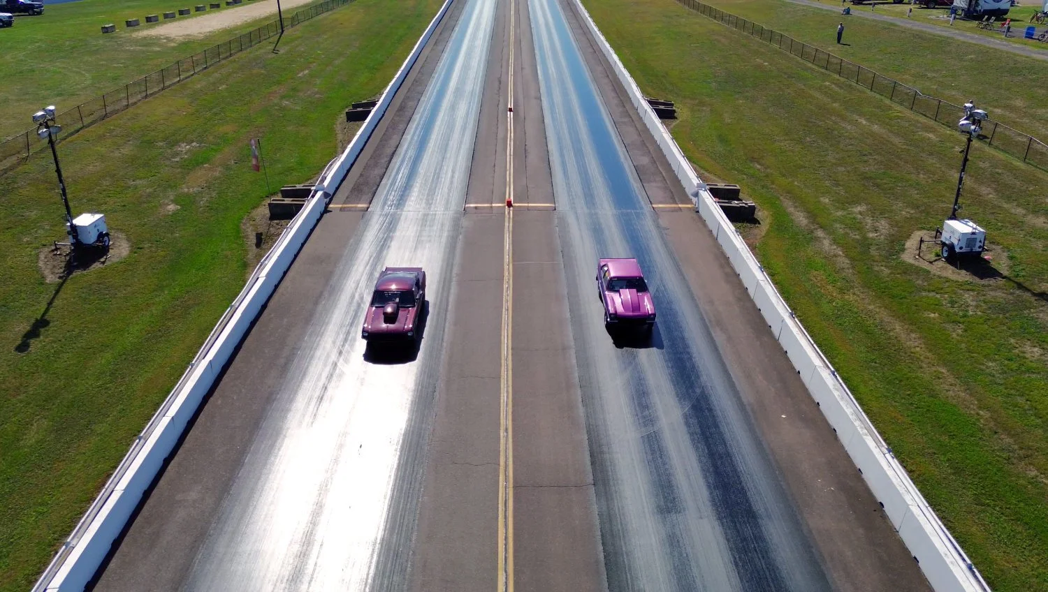 Aerial view of two racing cars on a drag strip, with the strip having a starting line and a strip of rubber down the center, surrounded by grassy areas and fencing.