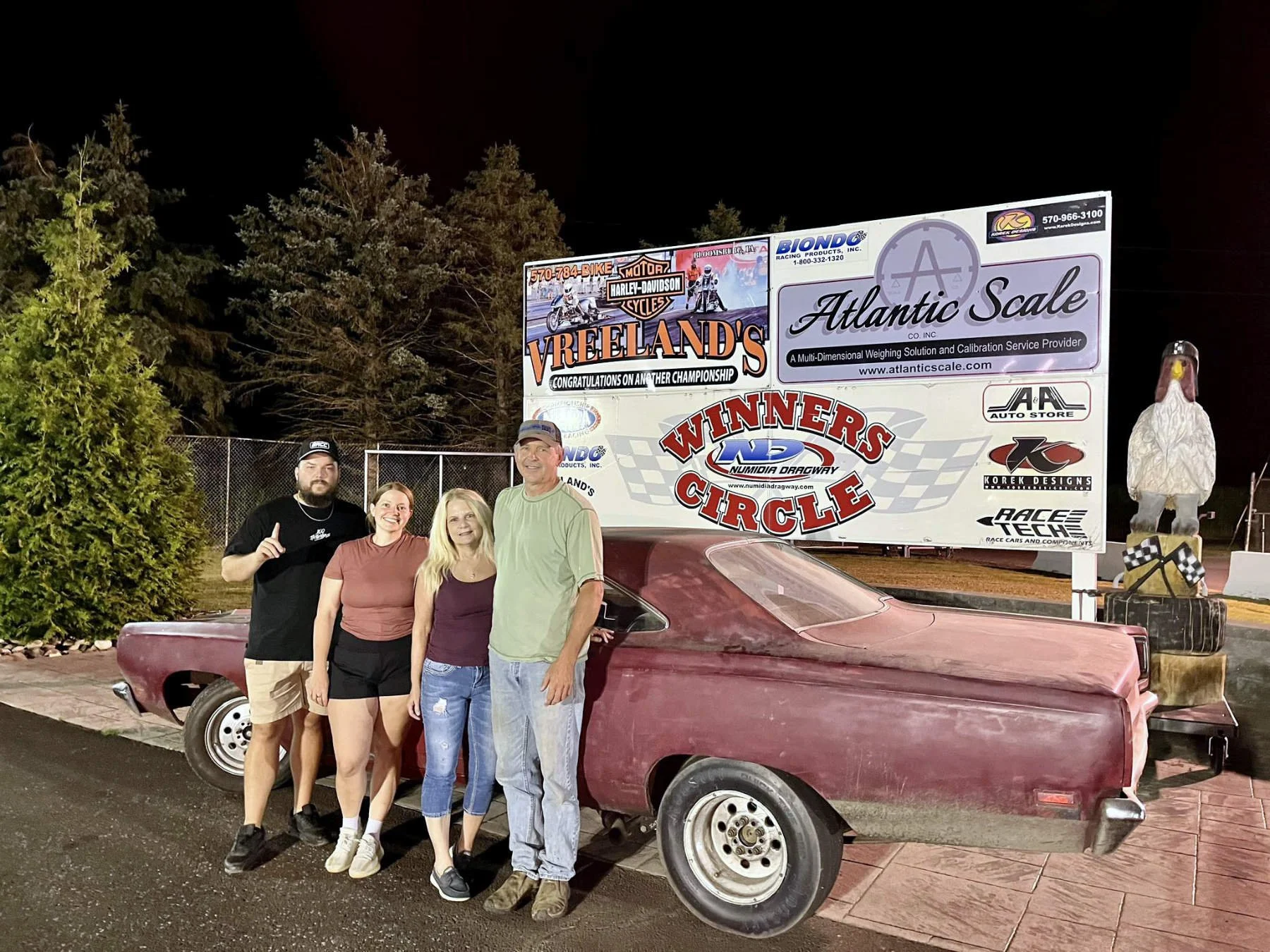 Four people standing in front of a vintage drag racing car at night, with banners and signs behind them including a statue of an eagle and various logos.