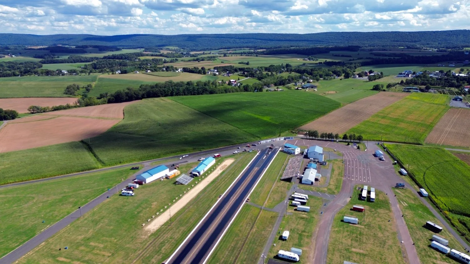 Aerial view of a rural area with farmland, roads, and buildings, showing fields, houses, and a highway with vehicles under partly cloudy skies.
