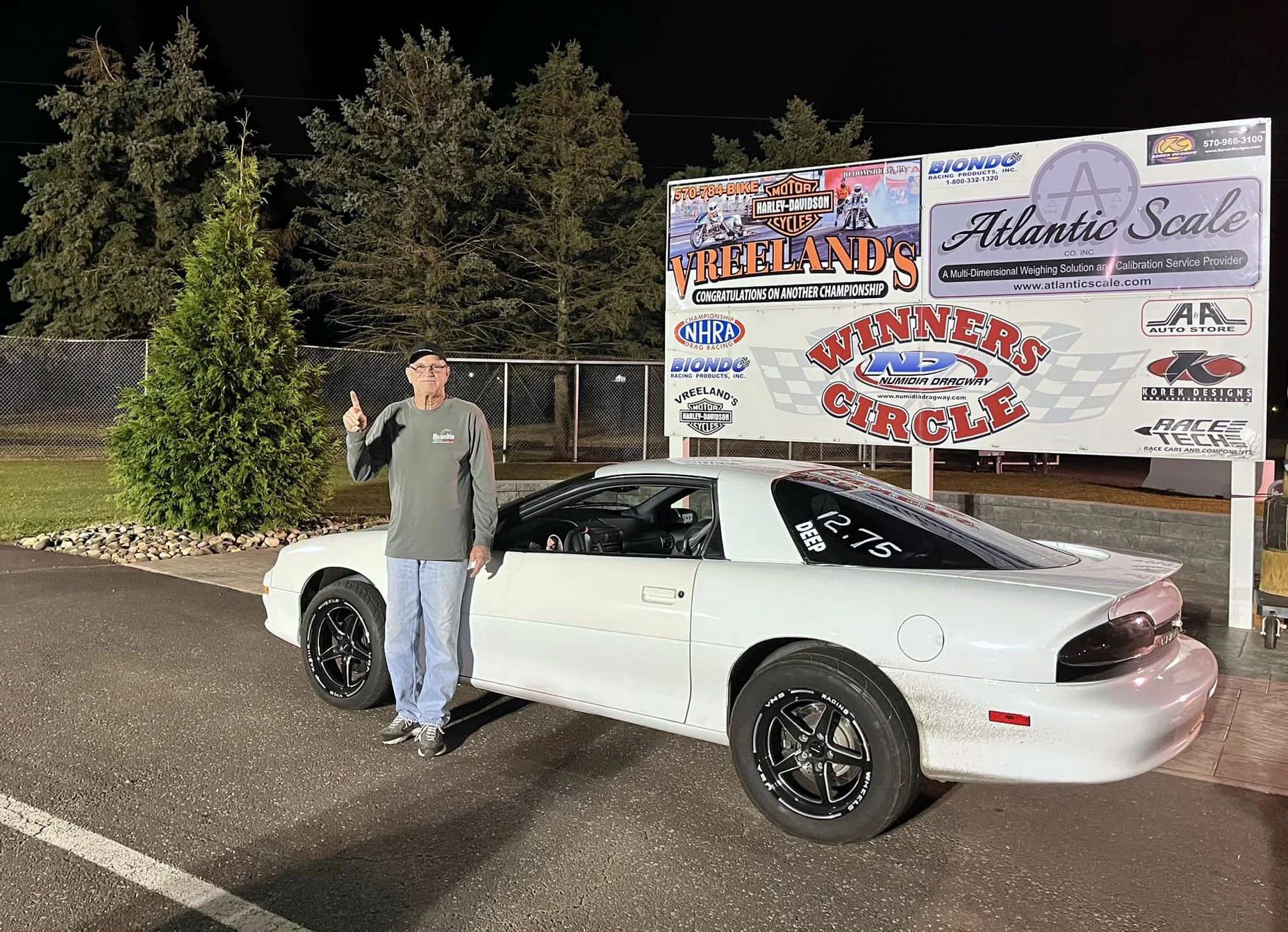 A man standing next to a white sports car with black wheels at night. Behind them, there is a large sign for Atlantic Scale and other sponsors, and tall pine trees in the background.