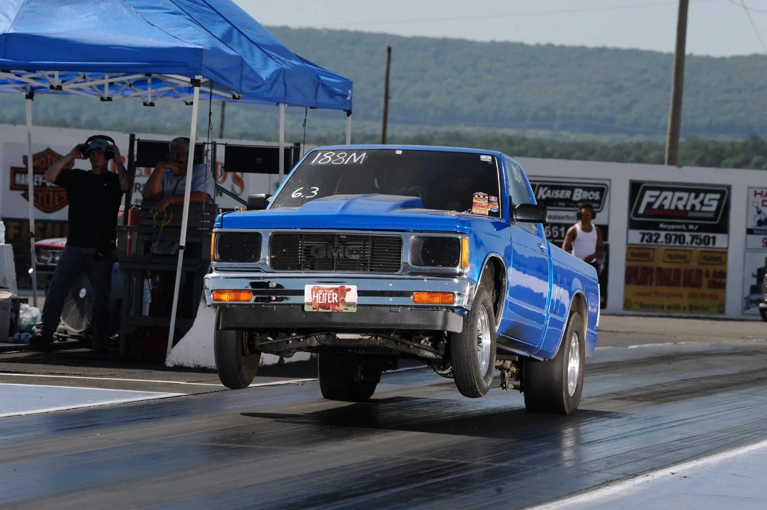 A blue vintage GMC truck with front wheels lifted off the ground on a drag racing strip, participating in a race, with two men standing under a blue canopy in the background and race banners visible.