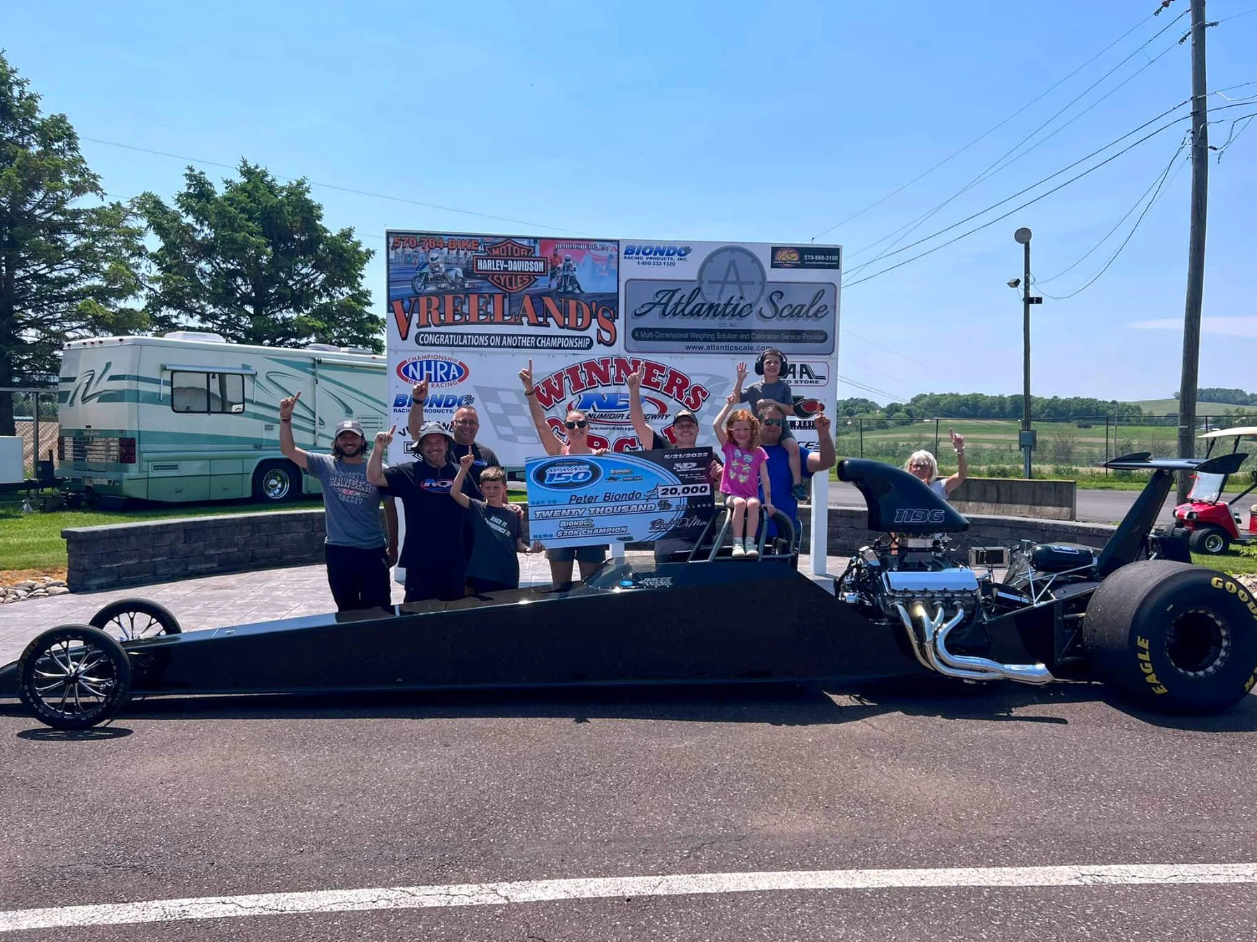 Group of people celebrating with a drag racing car and a large check at a racing event. They are on an outdoor track with banners and a RV in the background.