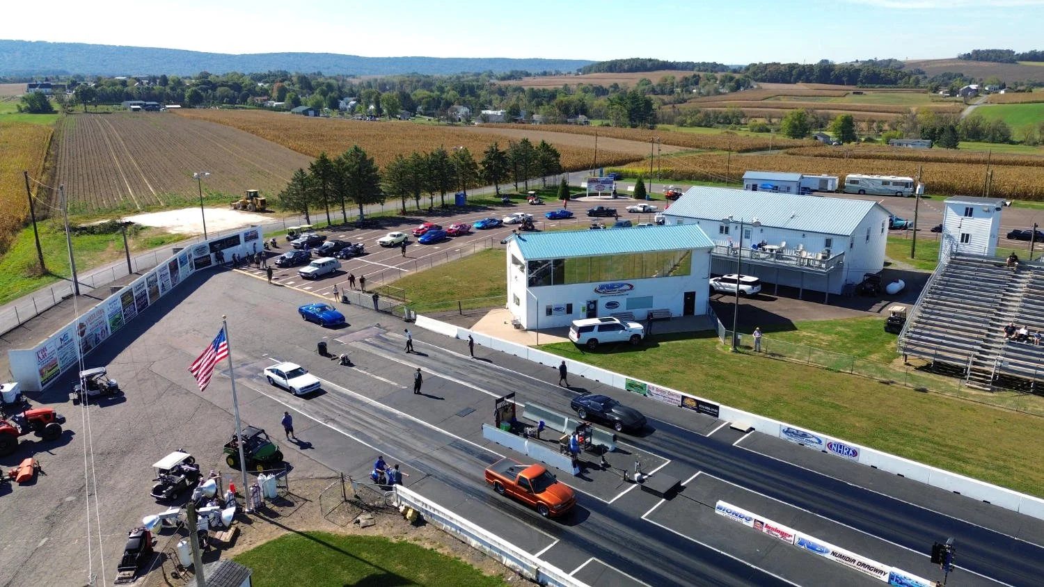 Aerial view of a drag racing track with cars and spectators, surrounded by grassy areas, buildings, and farmland in the background.