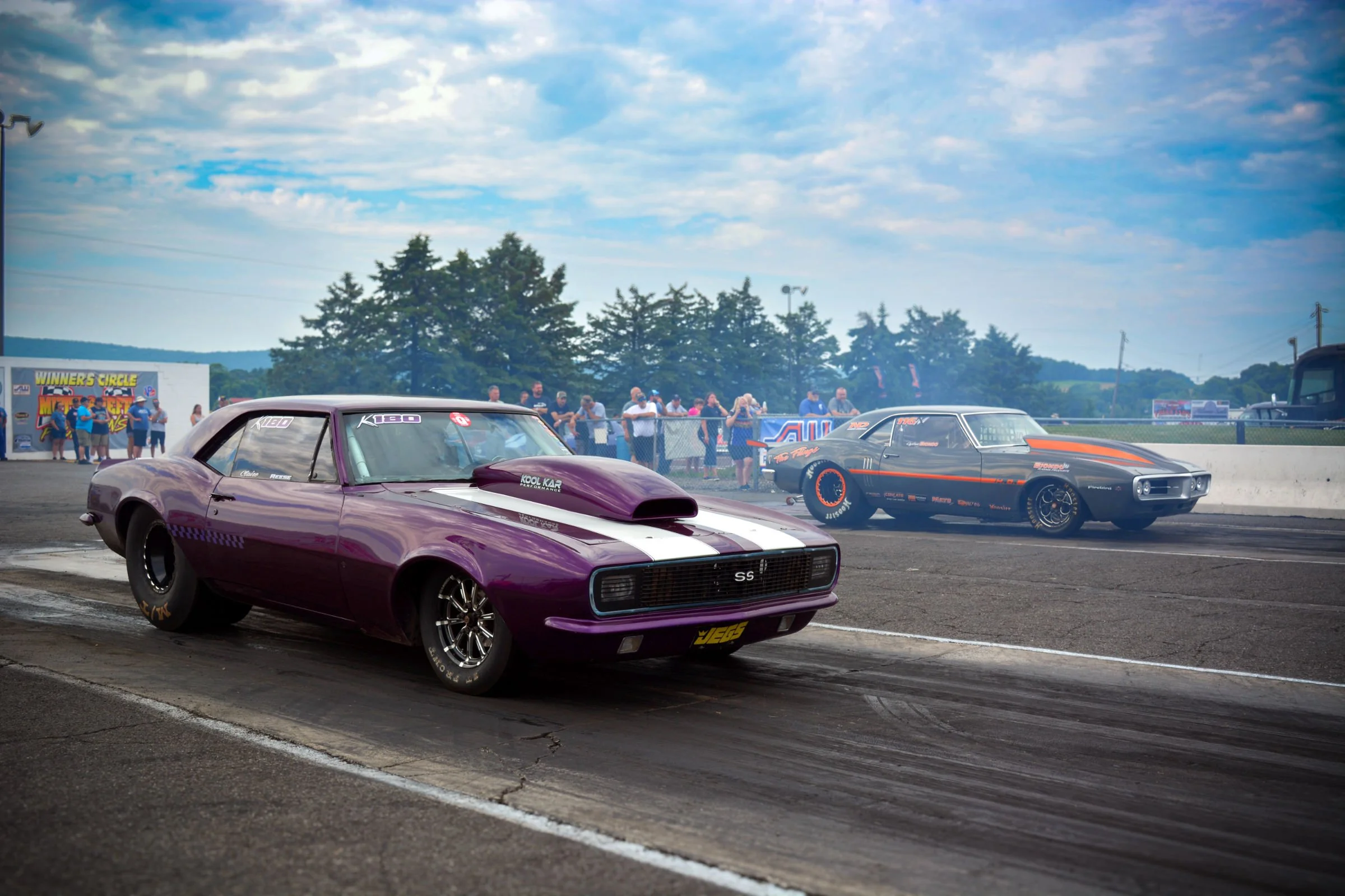 A drag racing event showing classic muscle cars on the starting line, with spectators watching behind a fence on a cloudy day.