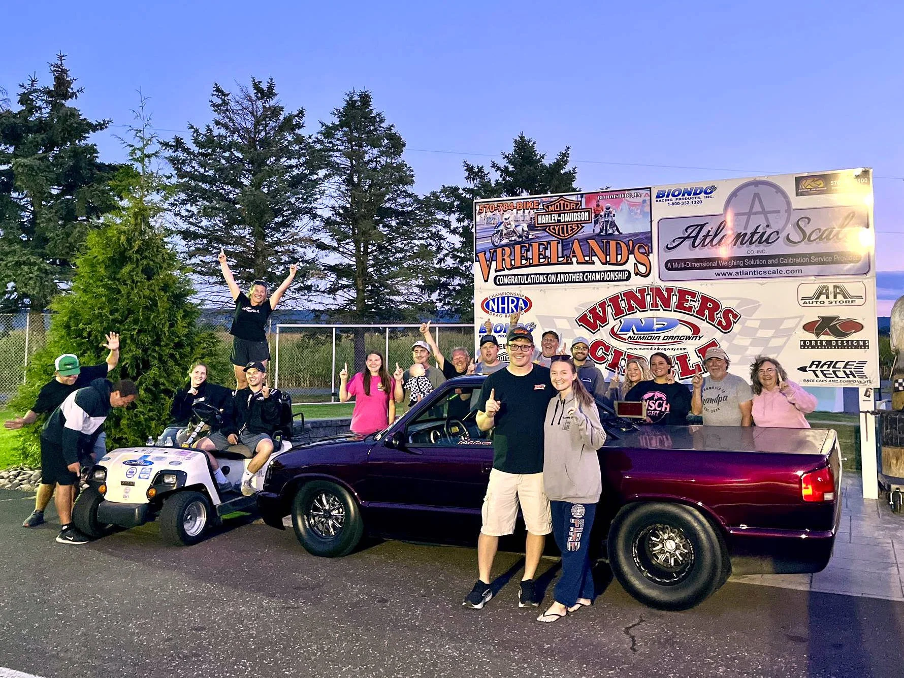 Group of people celebrating after winning at a drag racing event, posing in front of a banner with motorcycle and automotive sponsorships, with two golf carts and a black pickup truck, surrounded by trees and a fence, during early evening.