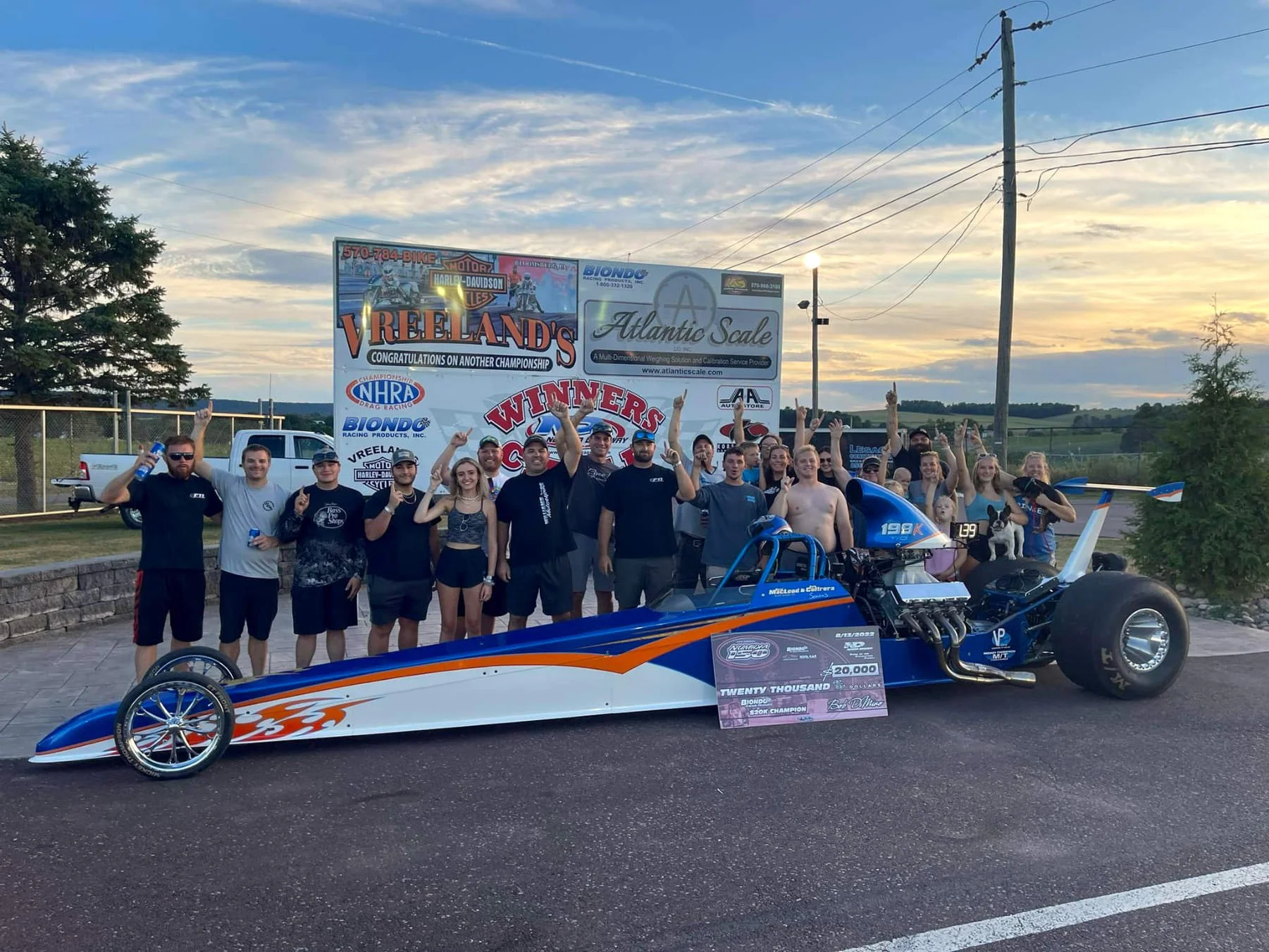 Group of people celebrating next to a drag racing car, holding a large check for $20,000 and a trophy, with banners and a race track in the background during sunset.
